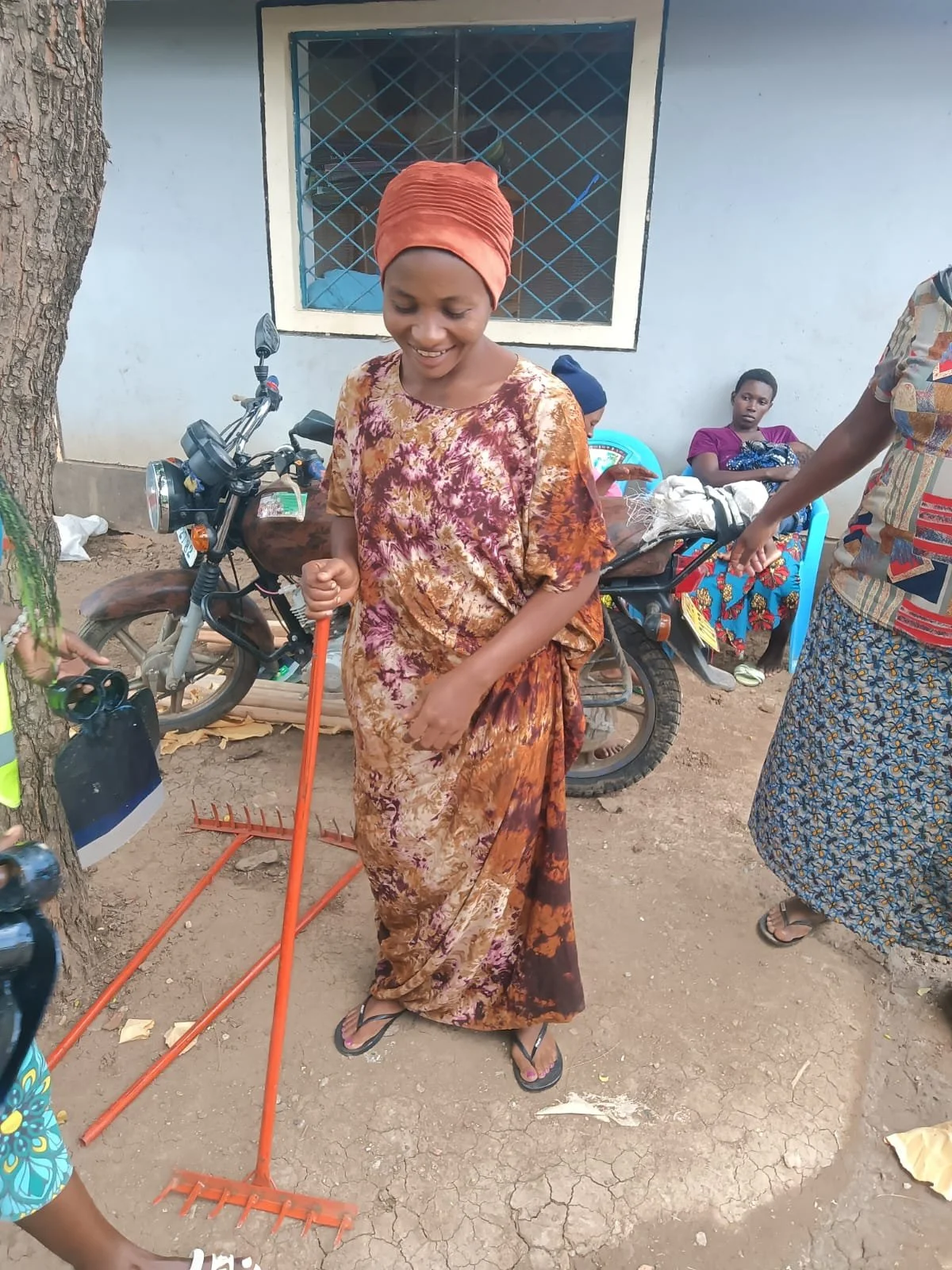 A woman in a brown and orange patterned dress and matching headscarf is smiling while holding a red rake. She is standing on dirt ground with a tree trunk on her left. In the background, there are two women sitting on a blue bench, beside a motorcycl