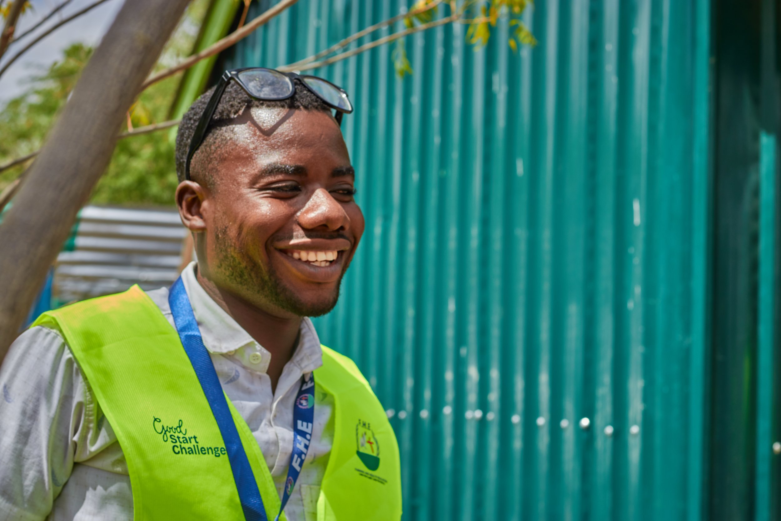 Smiling man with glasses on head wearing a high-visibility vest in front of a blue-green fence.