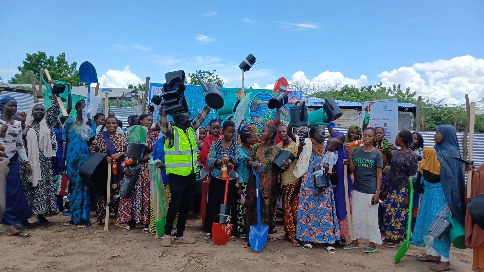 A crowd of people, mostly women, gathered outdoors under a partly cloudy sky, holding various tools and containers, with some raising their hands and smiling, possibly at a community event or distribution.
