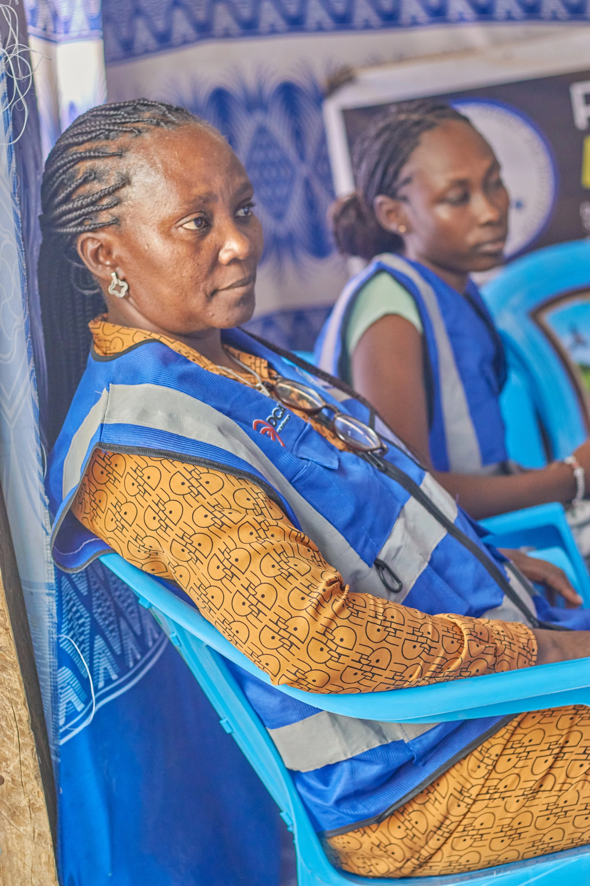 Two women sitting down dressed in matching blue uniforms, sitting on blue plastic chairs, at an event or gathering.