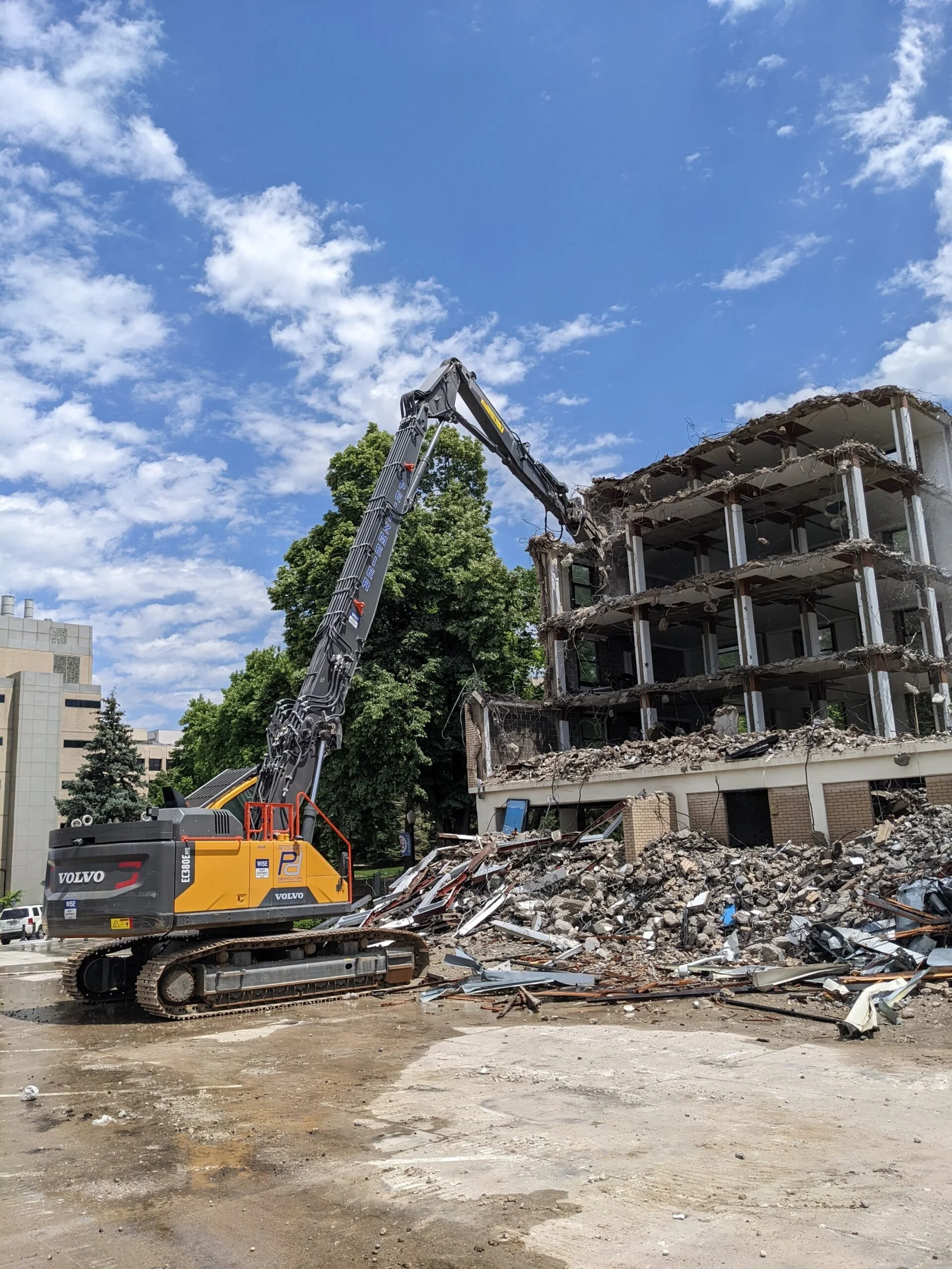 A construction excavator demolishing an old building with debris in the foreground, blue sky with clouds.