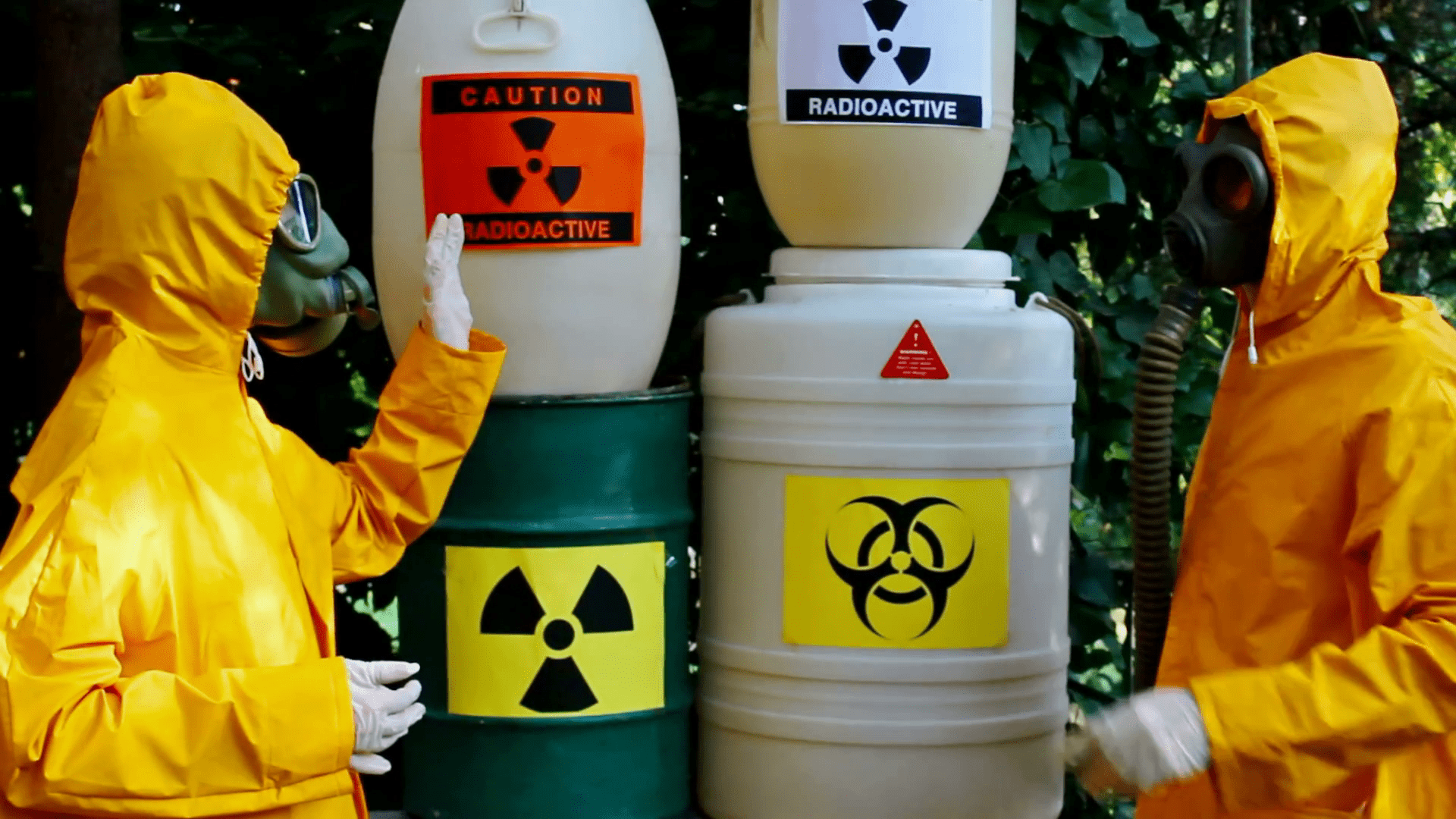 Two individuals in yellow hazmat suits with gas masks standing in front of barrels labeled with biohazard, radioactive, and caution signs.