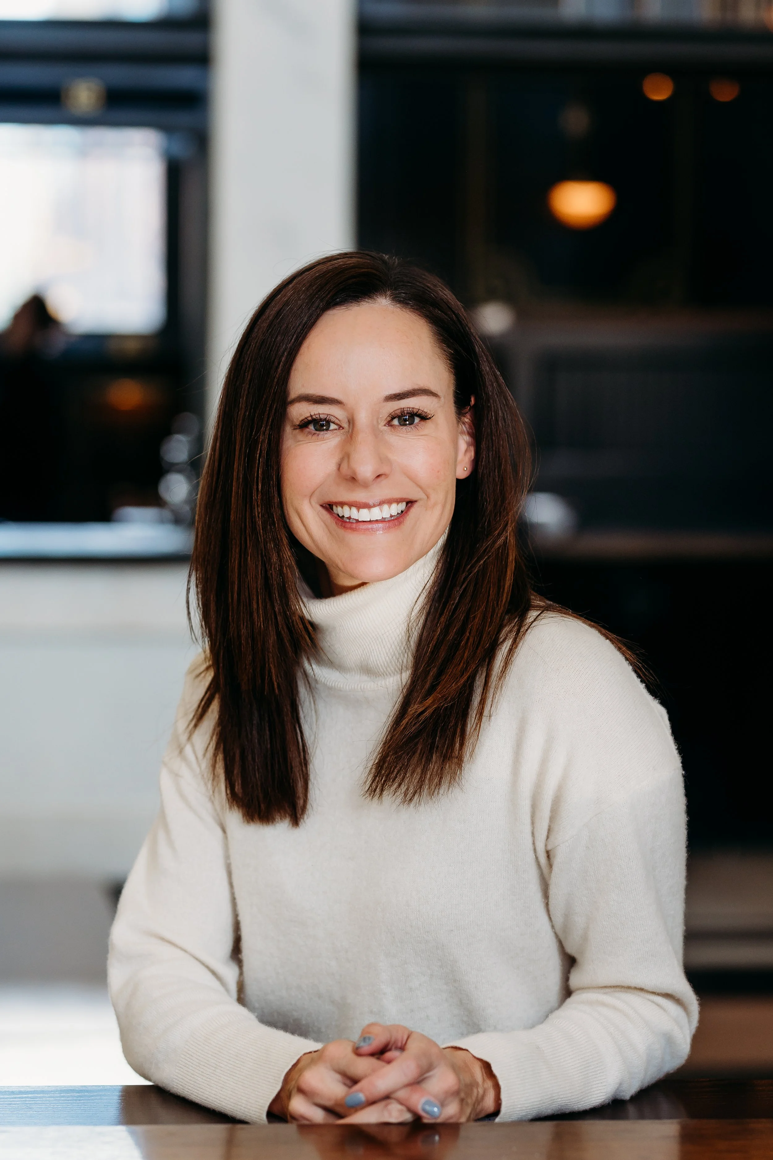 A woman with long brown hair wearing a cream turtleneck sweater, smiling while seated at a table in a modern indoor setting.