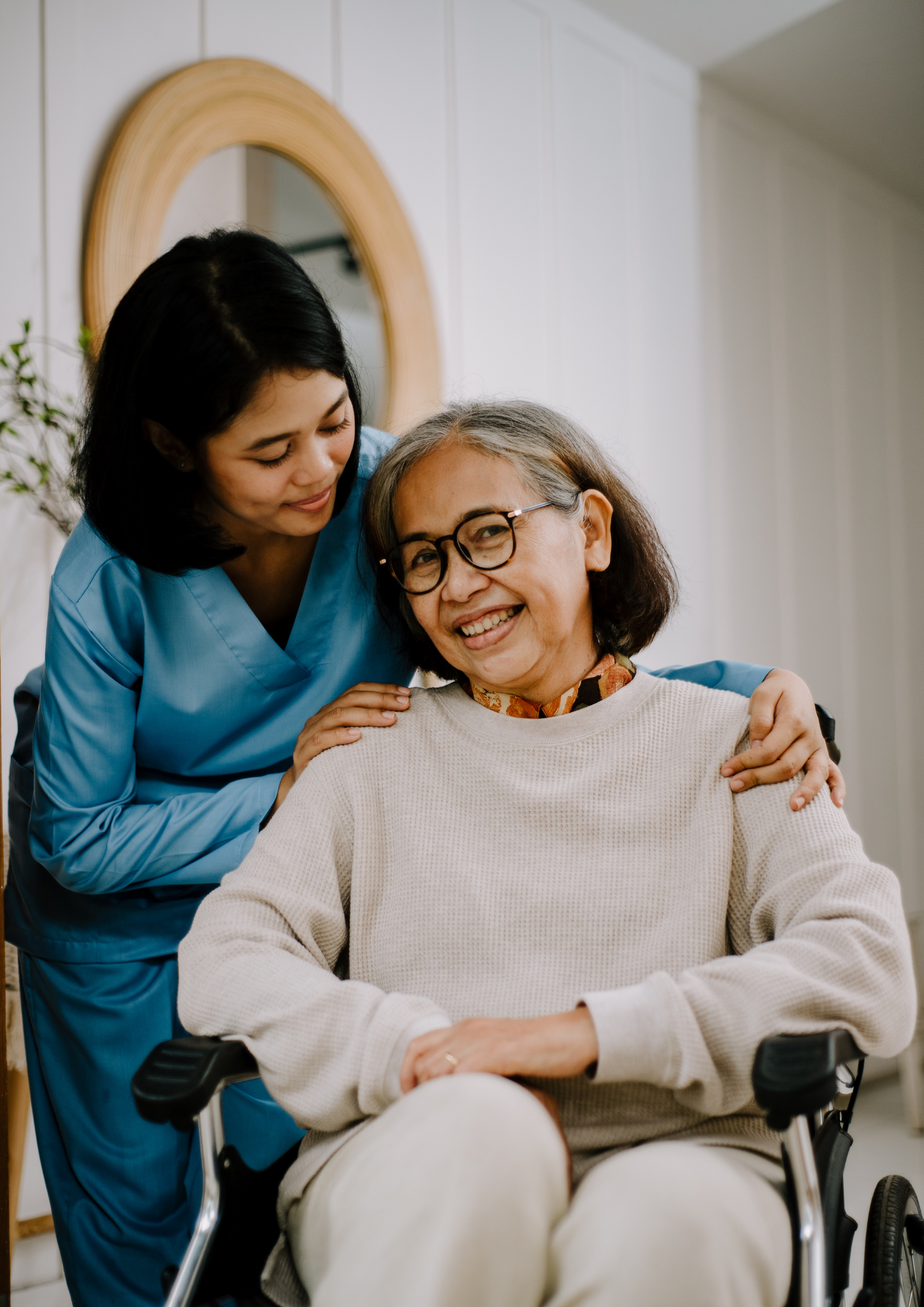 A caregiver in medical scrubs hugging an elderly woman in a wheelchair and smiling.