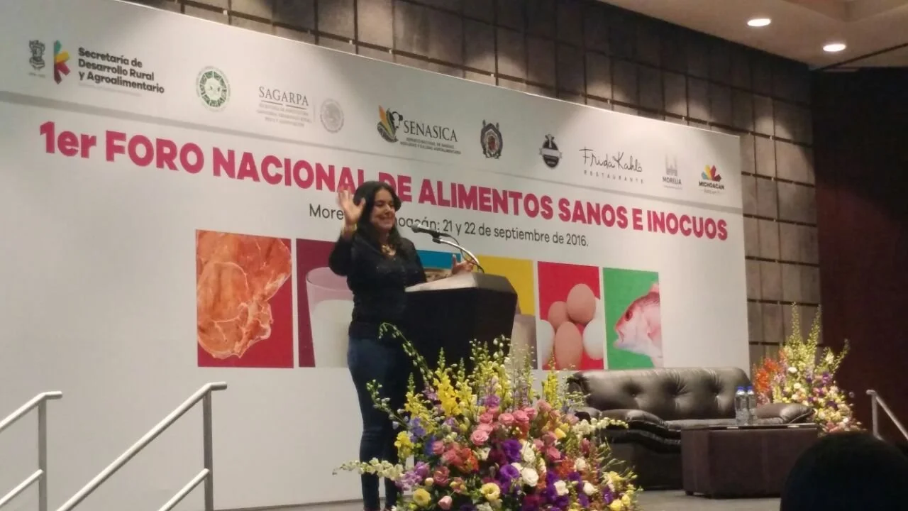 Una mujer en un escenario en un congreso o foro, con un podio y un fondo con logotipos y texto en español. La mujer está saludando y hay arreglos florales frente al escenario.