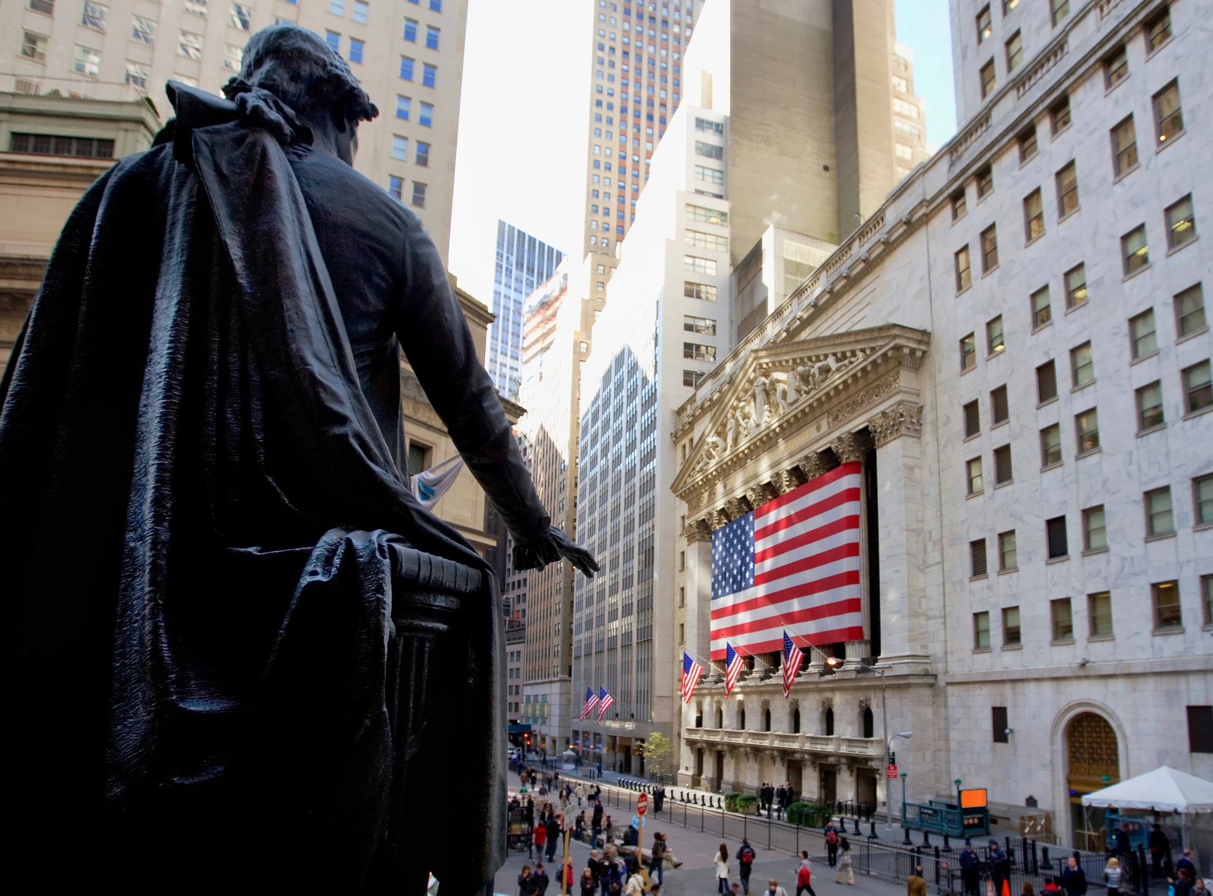 View from behind a seated statue of a historical figure overlooking a busy street scene in front of the New York Stock Exchange, decorated with American flags and a large American flag hanging on the building facade.