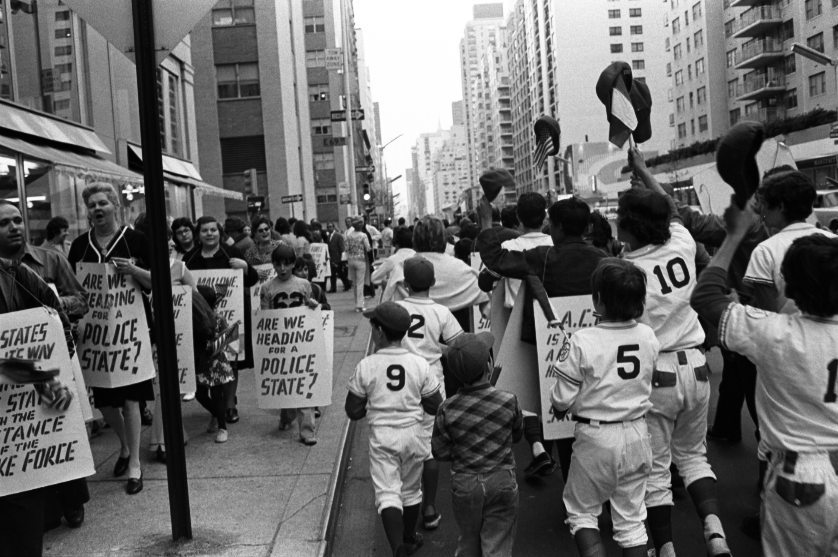 A black-and-white photo of a protest or rally with children and adults holding signs and banners on a city street with tall buildings.