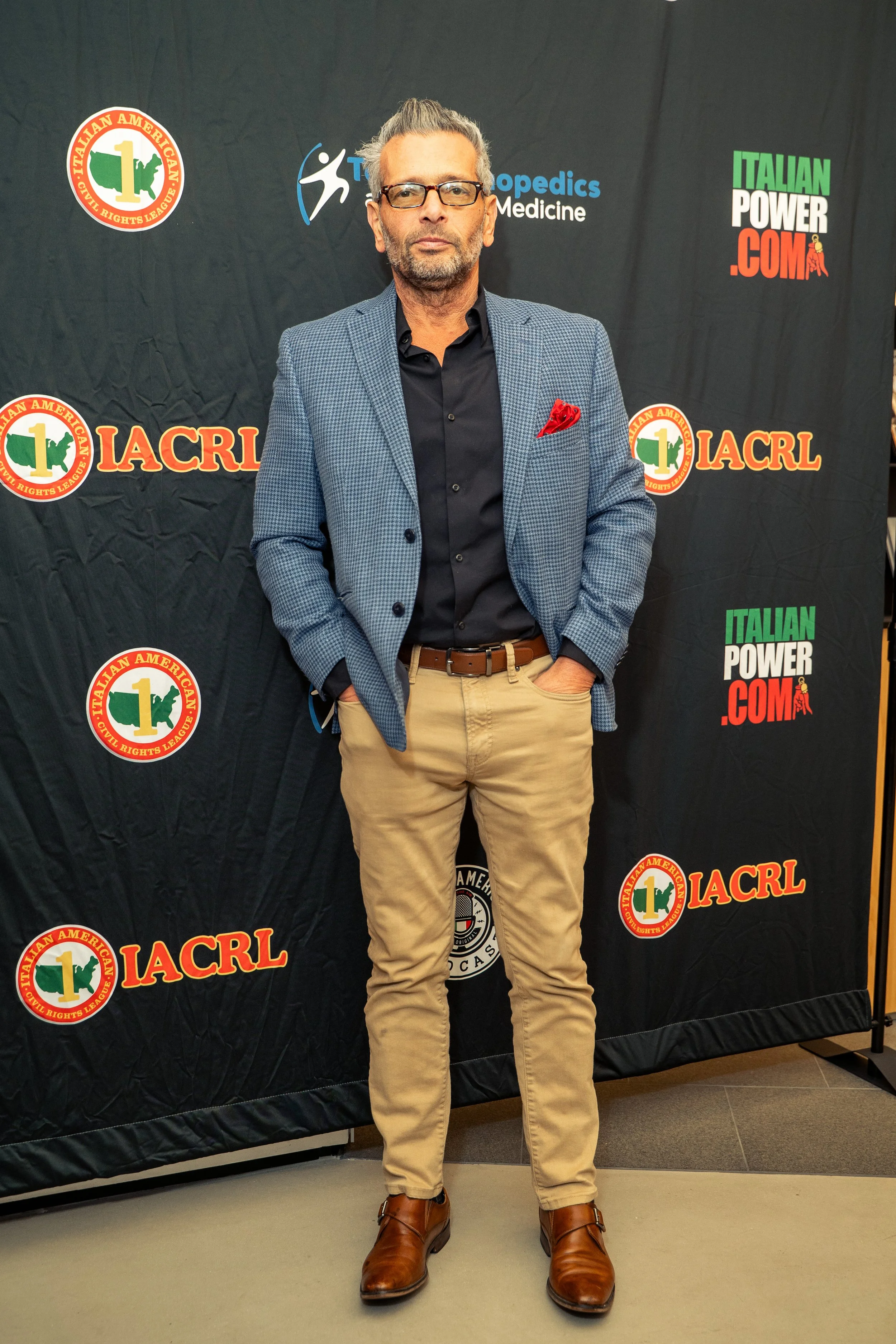 A man with gray hair, beard, and glasses standing with hands in his pockets in front of a black backdrop with logos for IACRL, Italian Power, and Orthopedics Medicine.