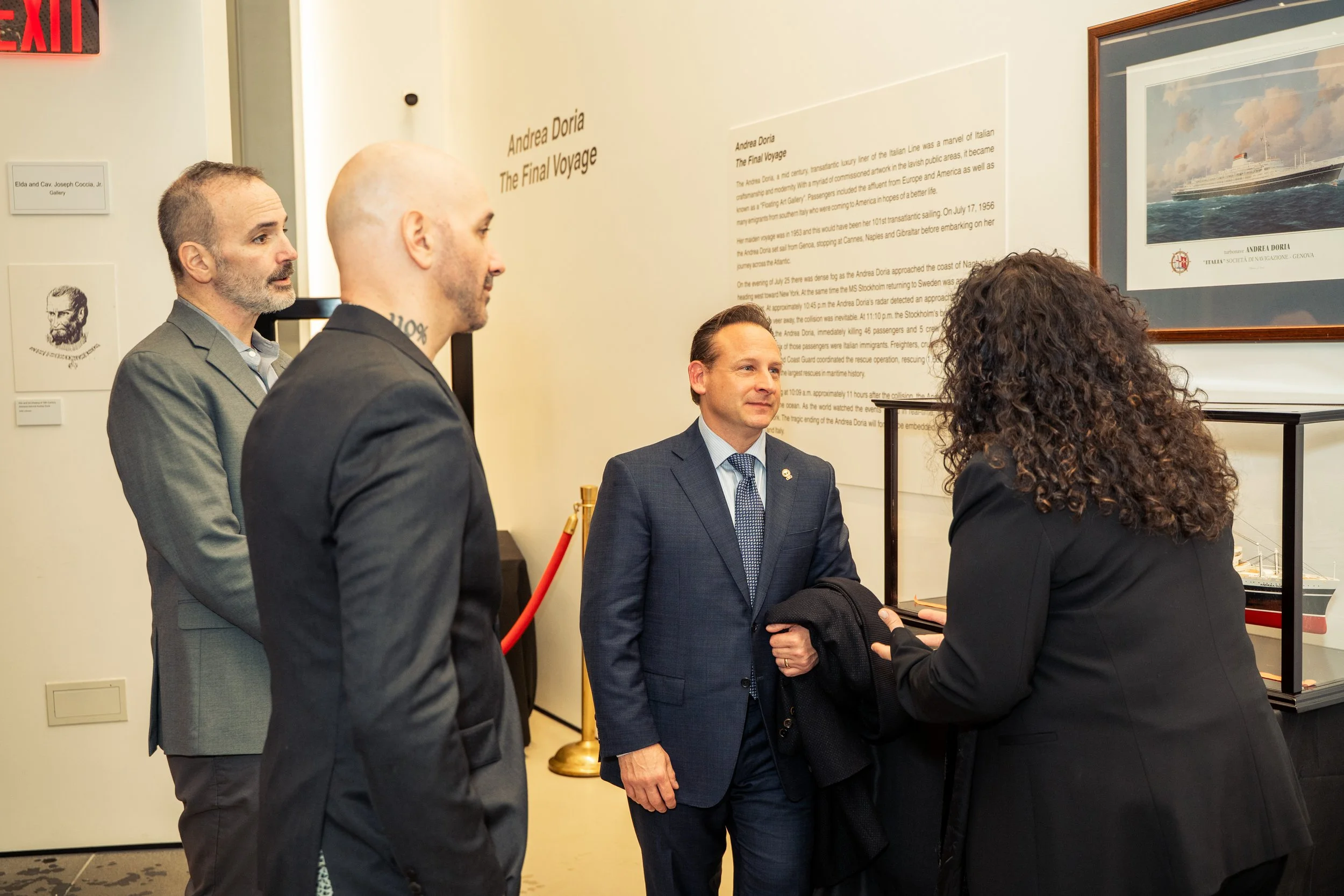 Four people standing and talking at an art exhibit or museum. One person is holding a coat while the others are listening. There are informational text panels and framed artwork on the wall in the background.