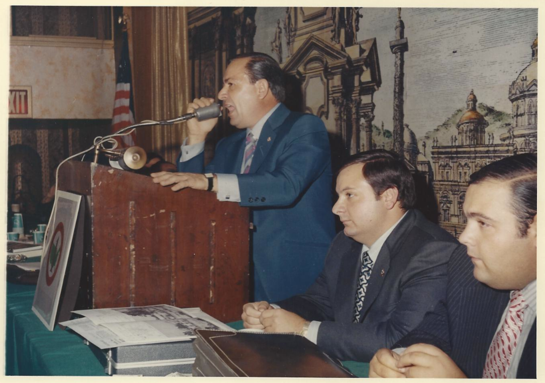 A man in a blue suit is speaking into a microphone at a podium during a formal event, with two men seated at a table beside him, all dressed in suits, a mural of buildings and an American flag in the background.
