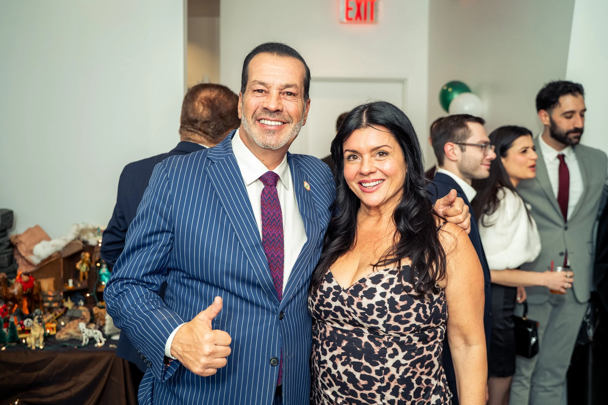 A man in a blue pinstripe suit and a woman in a leopard print dress standing together at an indoor event, smiling at the camera. Behind them, a group of people are engaged in conversation, with some holding drinks and festive decorations visible in t