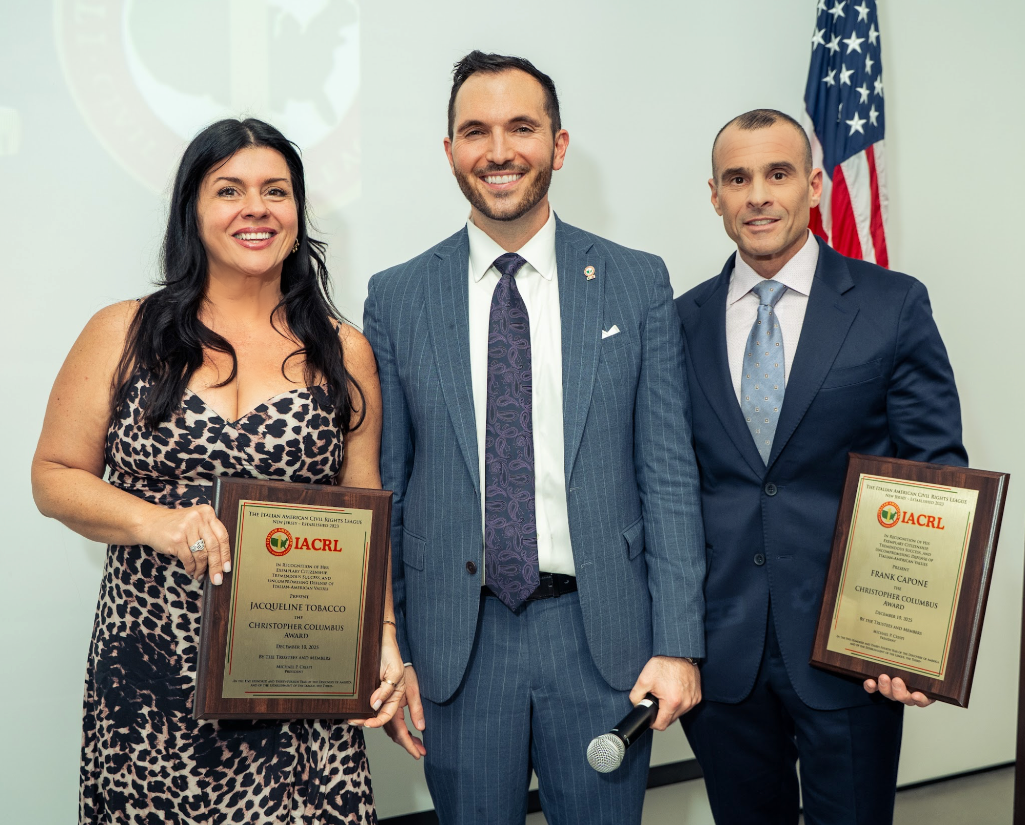 Three people standing in front of a white wall with an American flag. The woman on the left holds a plaque, the man in the middle holds a microphone, and the man on the right also holds a plaque. All are dressed in formal attire, smiling at the camer