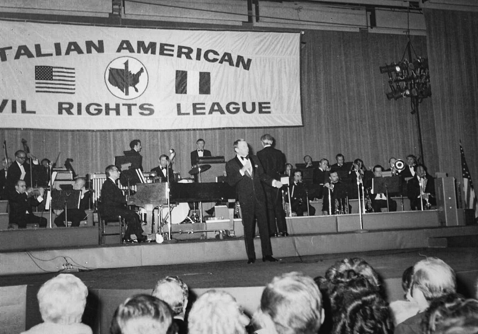 A black and white photo of a musical performance at an event for the Italian American Civil Rights League, with a large banner in the background, a stage with musicians, and a man speaking into a microphone.