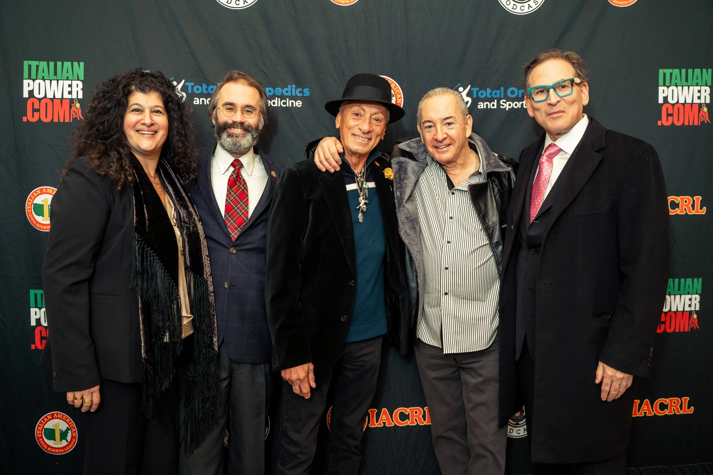 Group of six people standing together at an event, dressed in formal and semi-formal attire, with a backdrop displaying logos and text including 'Italian Power', 'Total Orthopedics', and 'LACRL'.