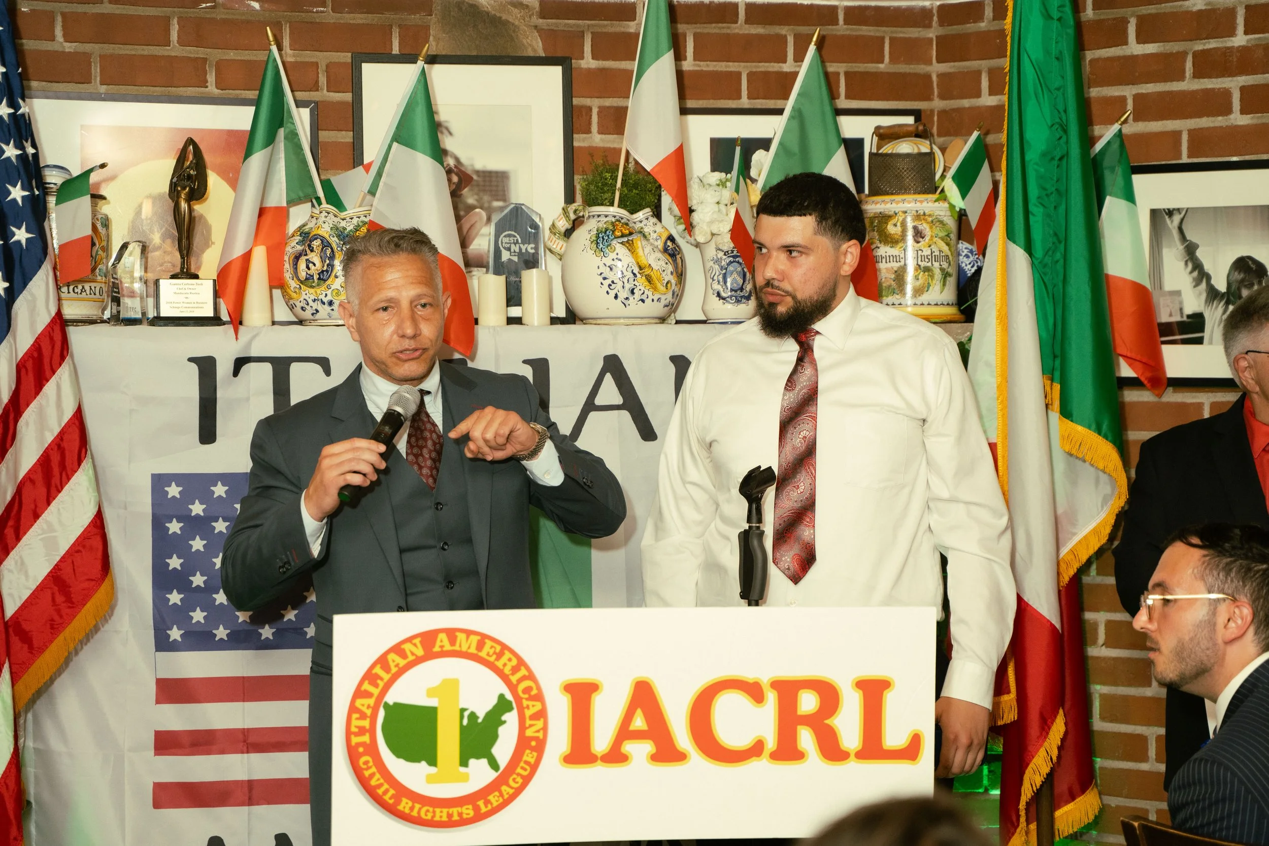A man with gray hair in a suit speaks into a microphone, standing next to a younger man with a beard in a white shirt and tie. They are at an event with multiple Italian and American flags, decorative vases, and framed pictures in the background. A b