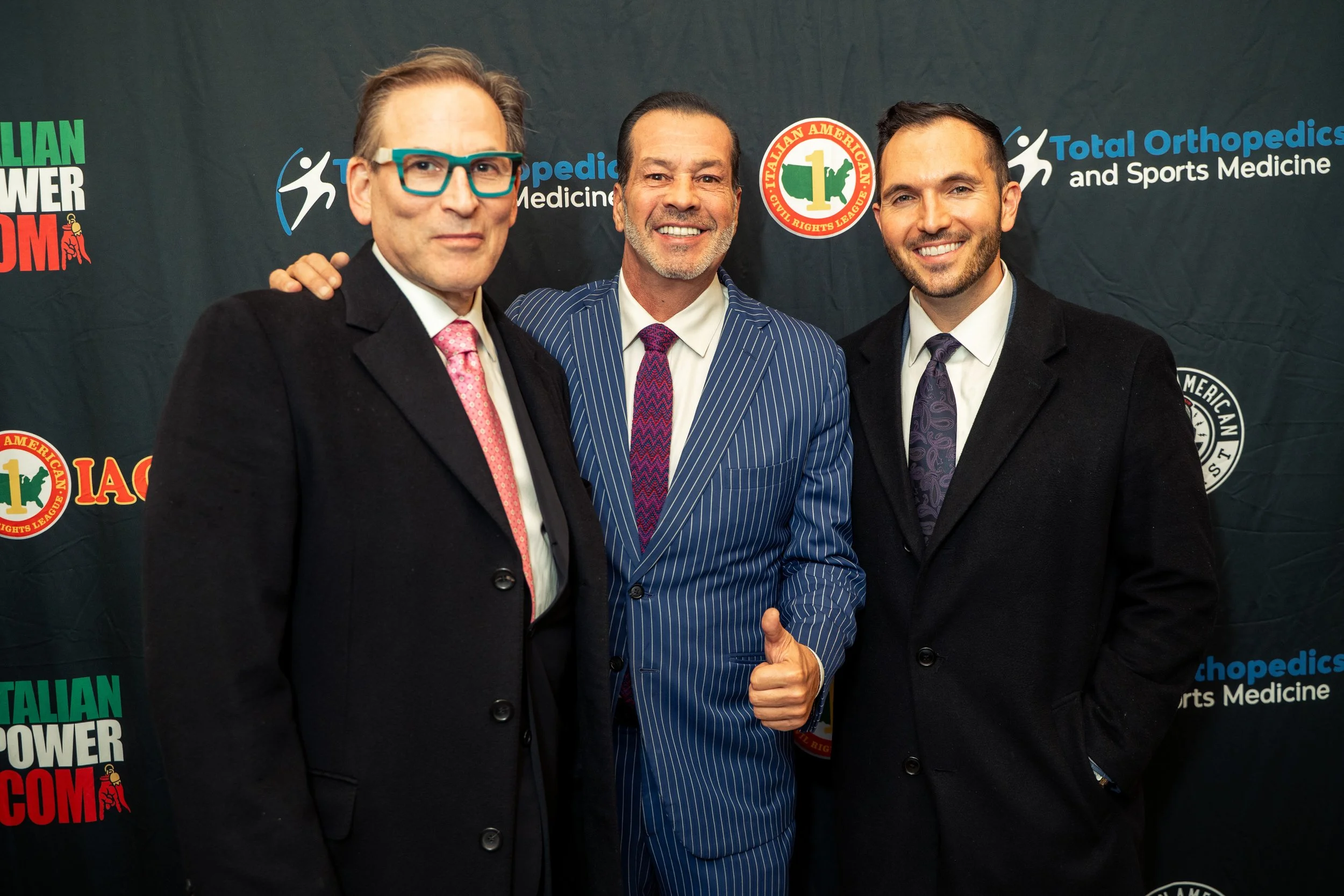 Three men in suits posing together at an event, with a backdrop featuring logos and text.