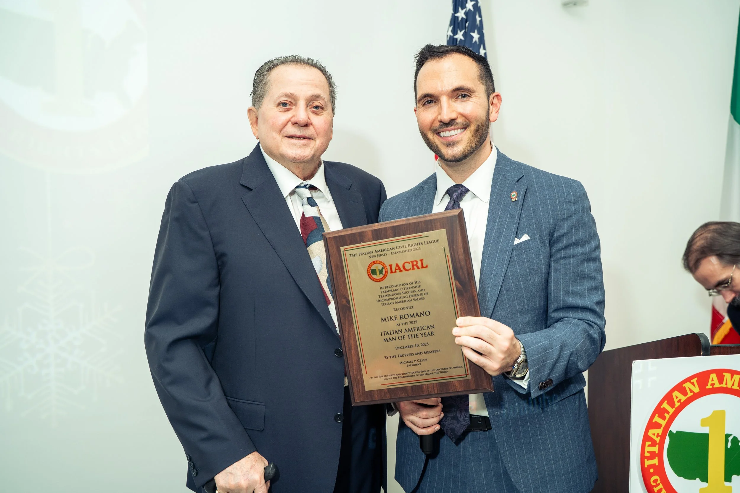 Two men in suits standing indoors, one holding a plaque with the logo 'IACRL' and text recognizing Mike Romano as Italian American Man of the Year, with an American flag in the background.