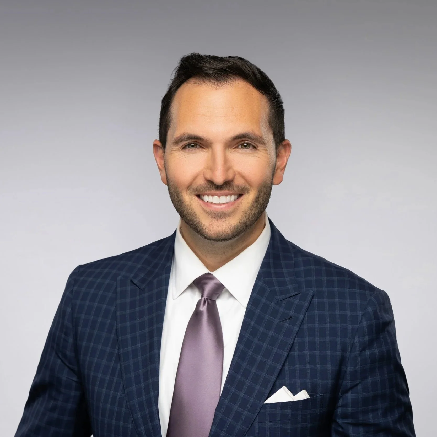 A man with dark hair and a beard wearing a suit and tie, smiling, with a gray background.