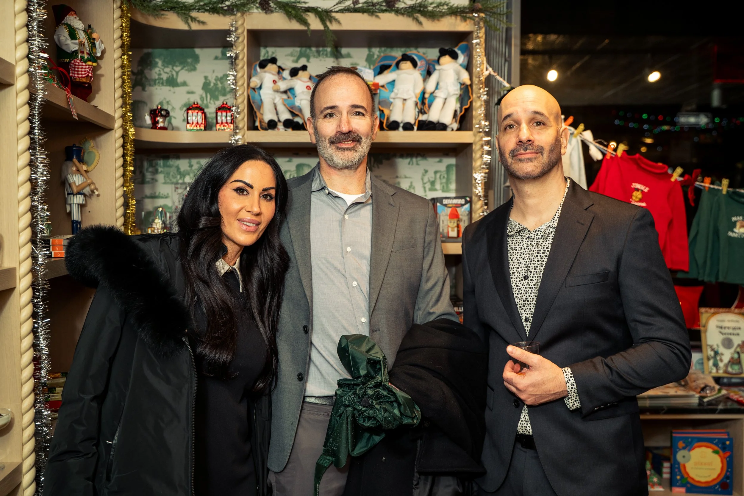 Three people dressed in formal attire standing in a Christmas-themed shop, with shelves of holiday decorations and plush toys in the background.