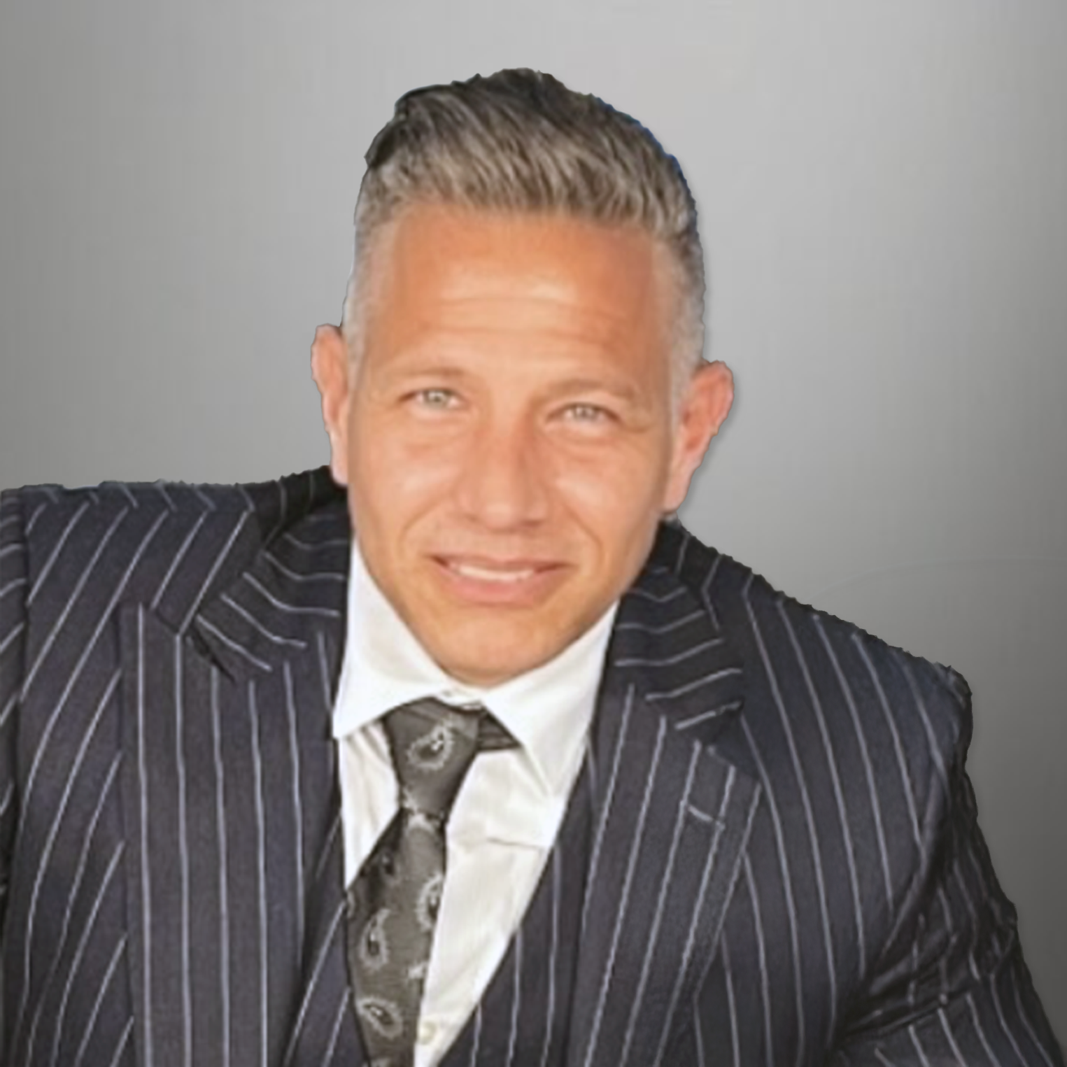 A man wearing a black pinstripe suit, white shirt, and a patterned tie, smiling at the camera, with a grey background.