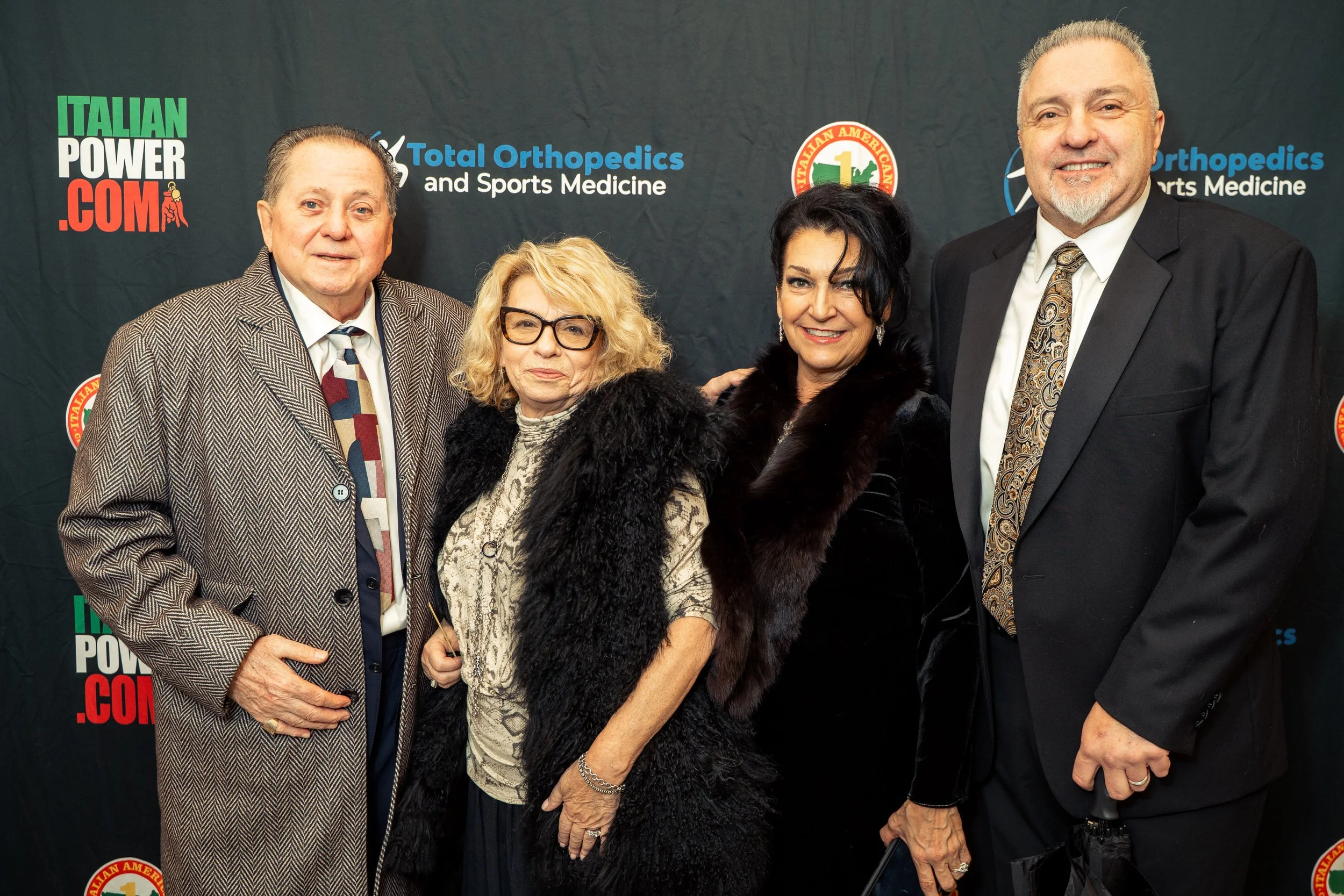 Four people standing in front of a backdrop with logos and text. The group includes two men and two women, all dressed in formal attire, with the women wearing dark, fur-lined coats.