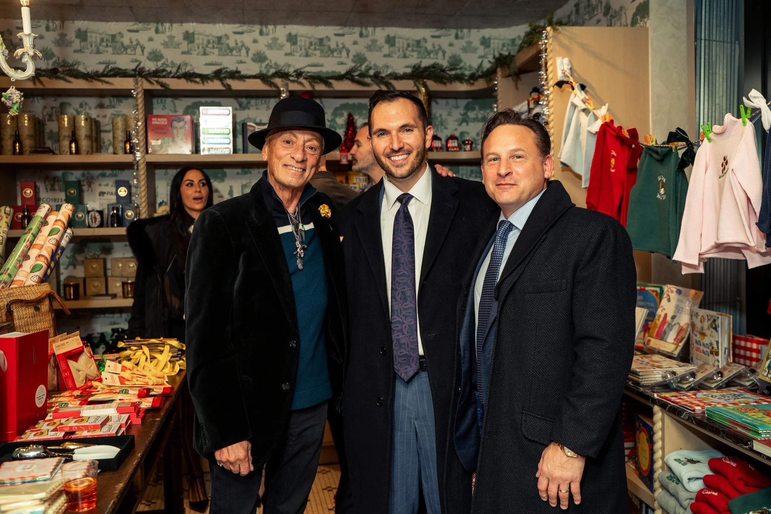 Three men in suits and holiday attire posing inside a festive store with Christmas decorations, wrapping paper, and holiday-themed merchandise.