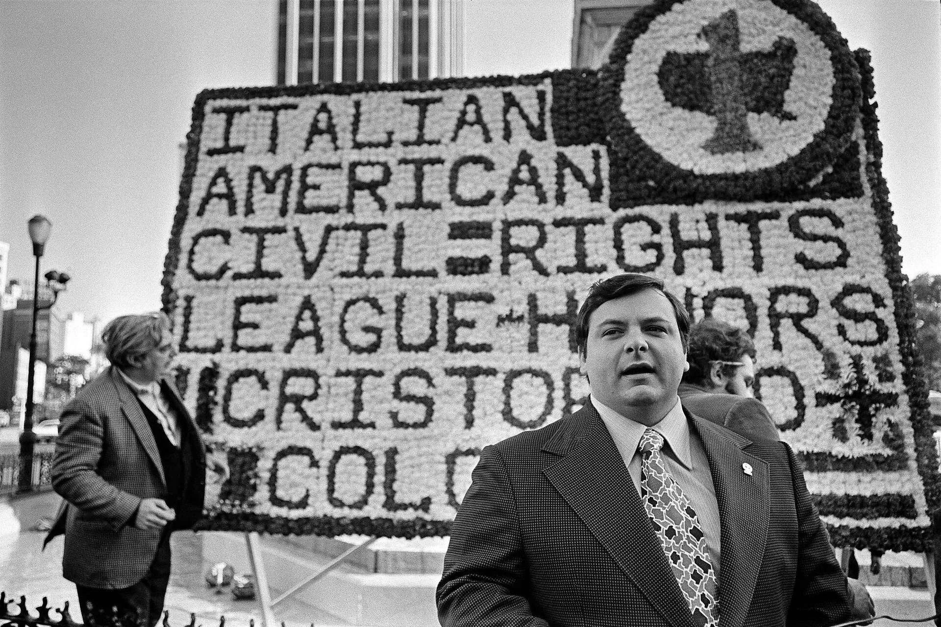 A man in a suit standing in front of a large floral tribute with messages supporting civil rights and equality, with two people and city buildings in the background.