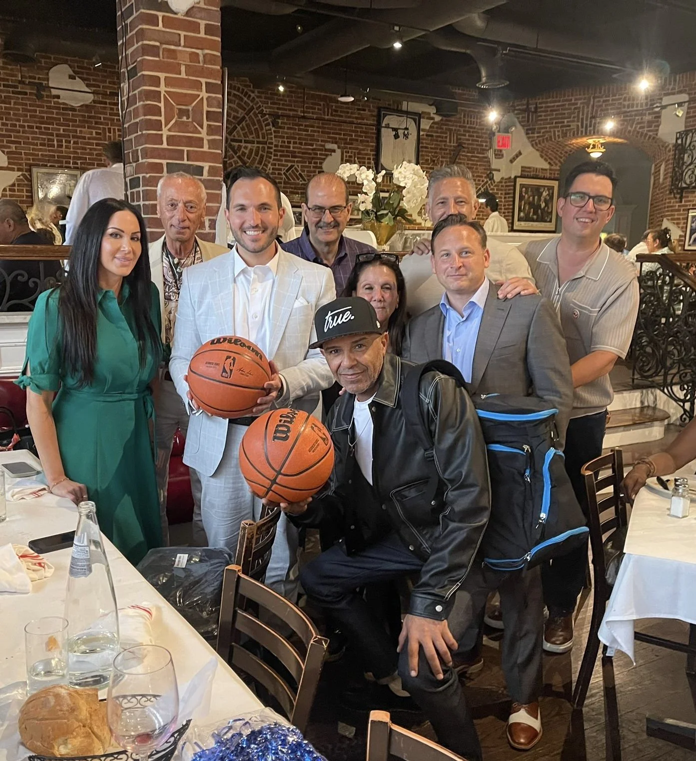 Group of nine people gathered in a restaurant, with two holding basketballs. The group includes men and women dressed in formal and casual attire, with some smiling at the camera. The setting features brick walls, framed pictures, and white flower arrangements.