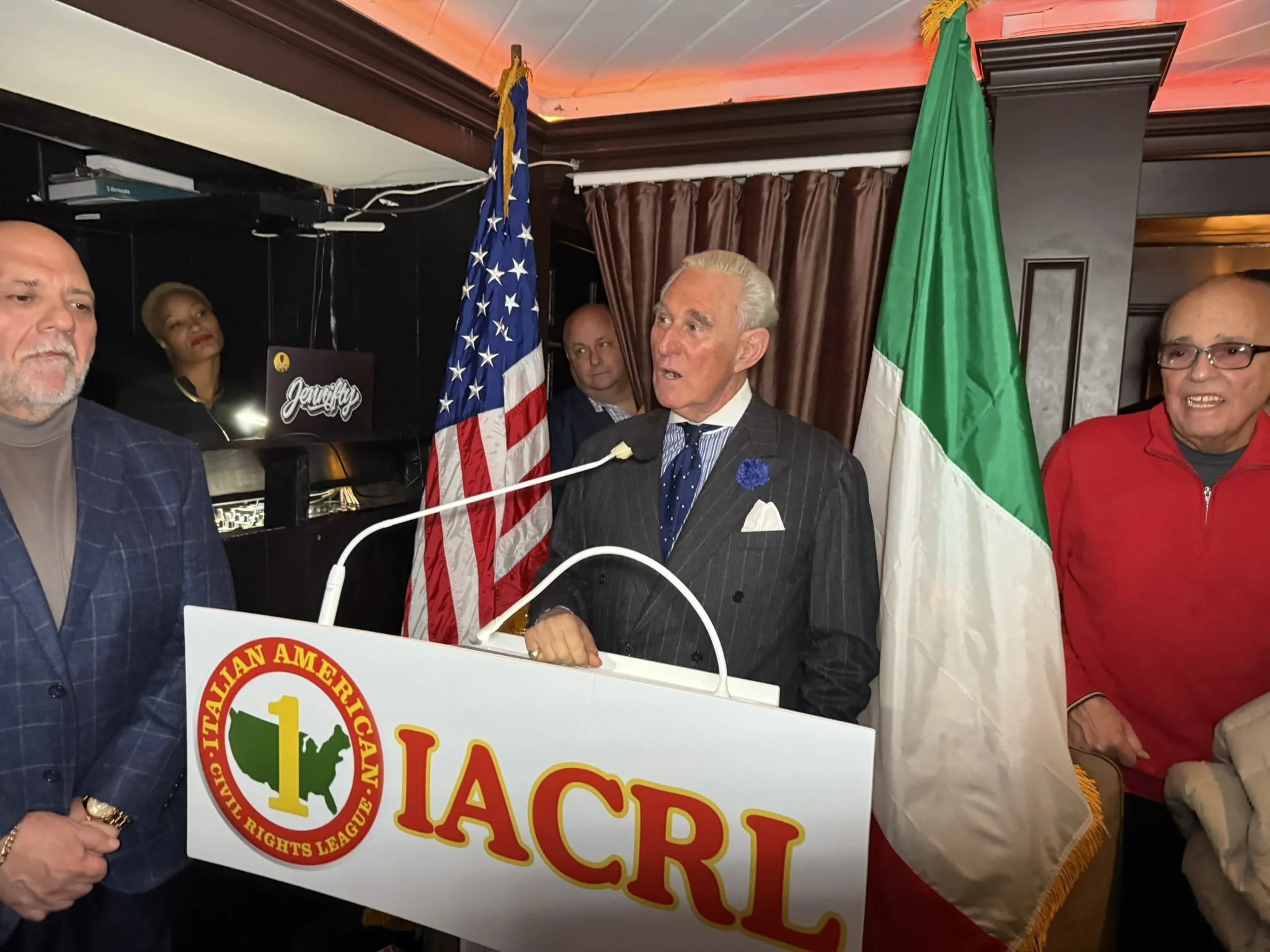 A man speaking at a podium with a sign that reads 'IACRL' and '1', flanked by the U.S. and Italian flags, with several people standing around him in a room decorated with dark walls and curtains.