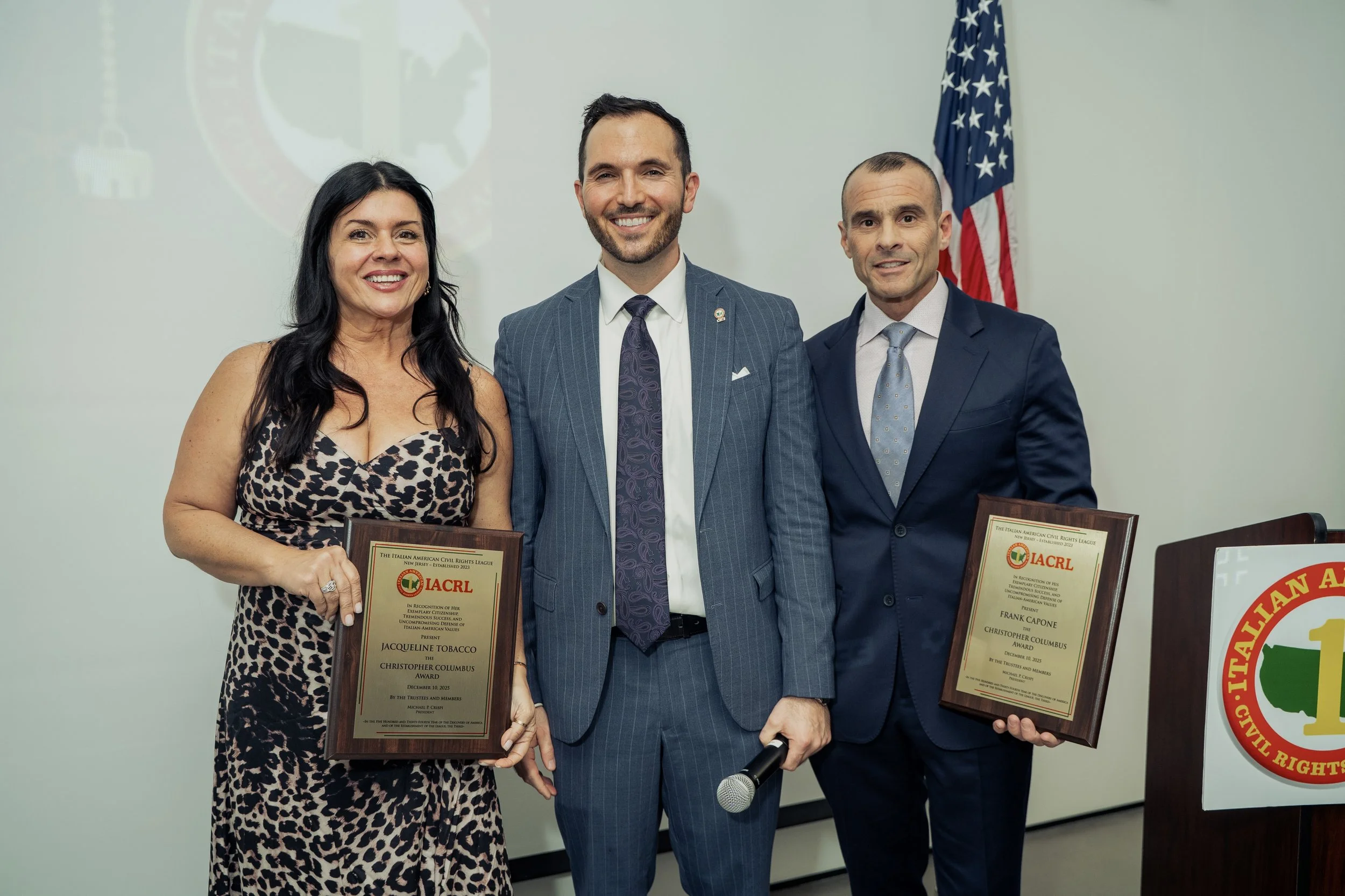Three people standing indoors, two men and one woman, smiling. The woman is holding a plaque, and the man on the right is holding another plaque. The plaques are awards from the Italian American Civil Rights League. An American flag is visible in the