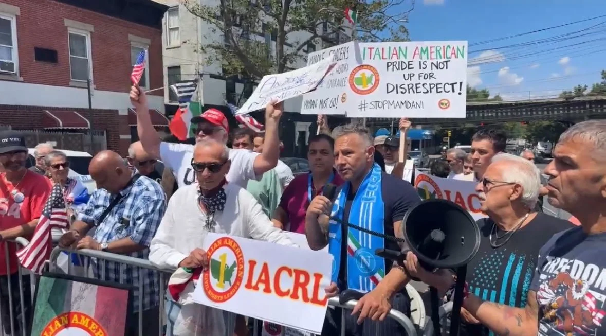 A group of protesters gathered outdoors holding signs and flags during a demonstration. Some signs say "Italian American pride is not up for disrespect" and "#stopmandani." The people appear to be advocating for Italian American pride and rights, with some wearing patriotic attire and using a megaphone.