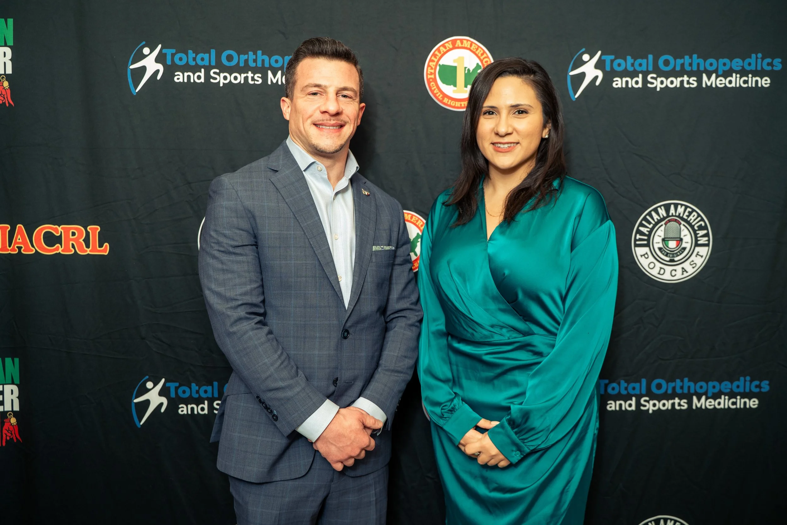 A man and a woman standing together in front of a backdrop with logos for Total Orthopedics and Sports Medicine, Italian American Podcast, and other organizations. Both are smiling and dressed in business attire.