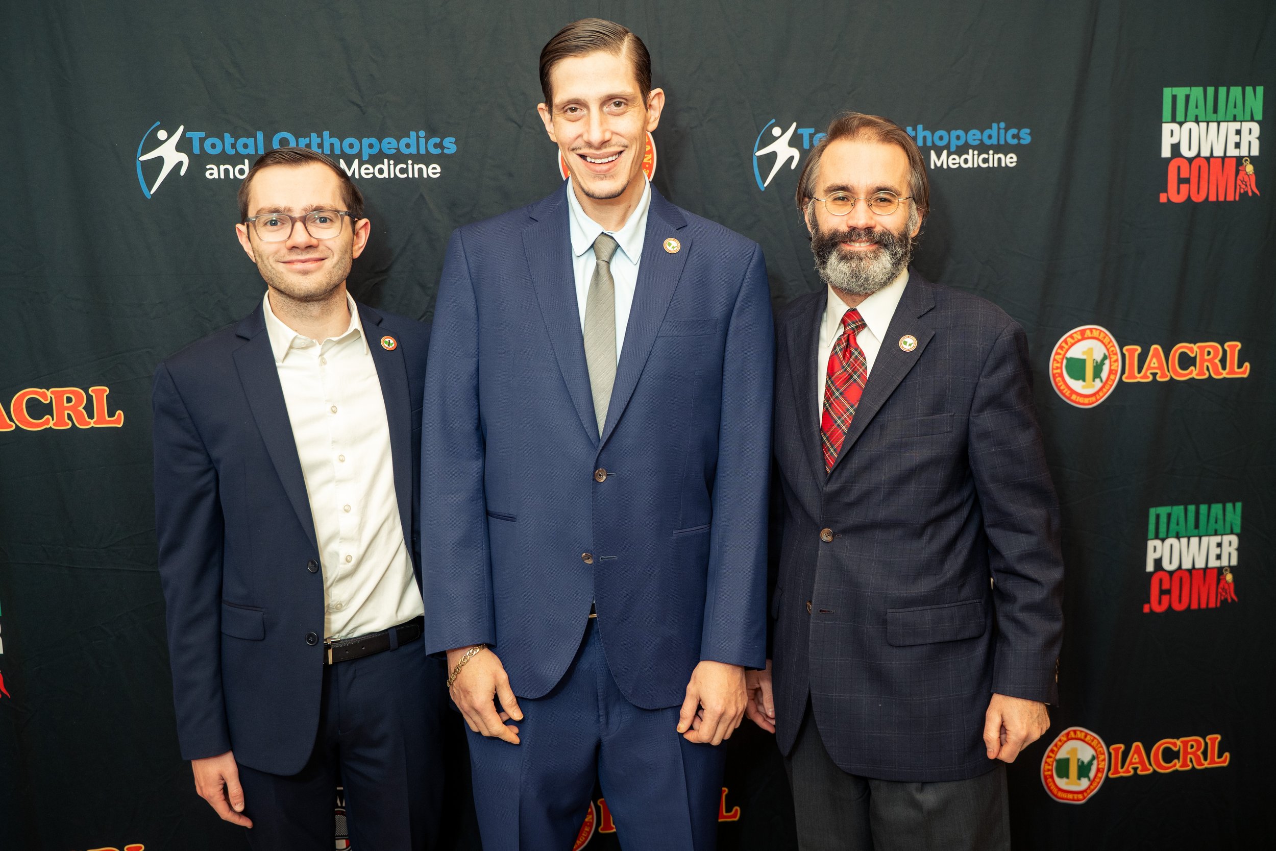 Three men in suits standing in front of a backdrop with logos for Total Orthopedics and Medicine, Italian Power, and LACRL, smiling at the camera.