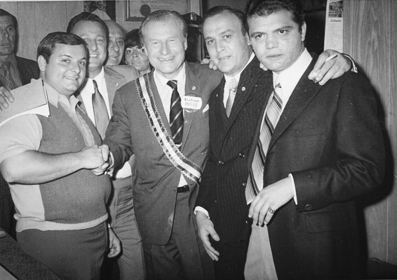 Black and white photo of a group of men dressed in suits, standing together indoors, smiling, with some showing a handshake.