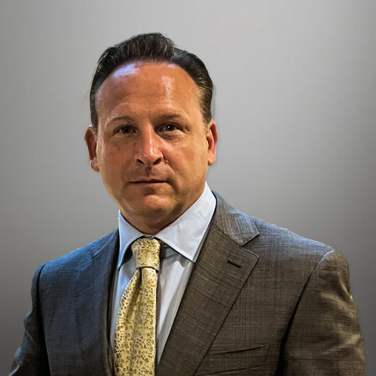 A man in a suit with a white shirt and a yellow patterned tie, standing against a plain gray background.