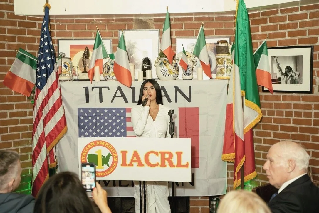 Woman speaking at a podium with flags of Italy and the USA, and a banner with 'ITALIAN' and 'IACRL' on it, surrounded by flags and decorations, in front of brick wall.