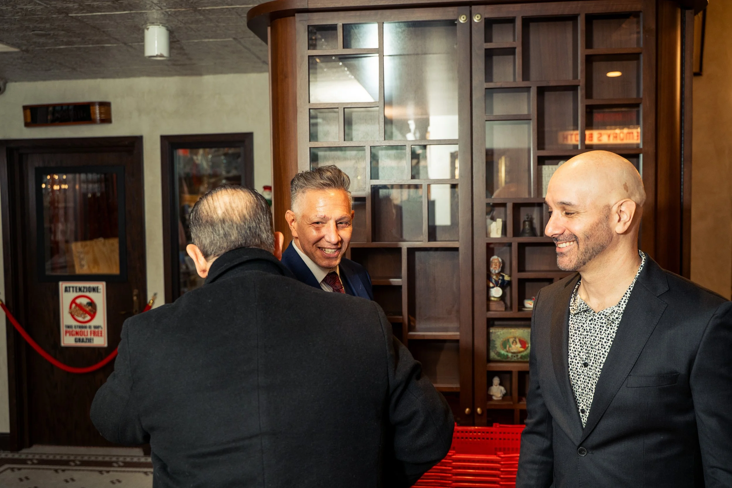 Three men in suits engaged in conversation inside a restaurant or bar, with wooden decor and shelves in the background.
