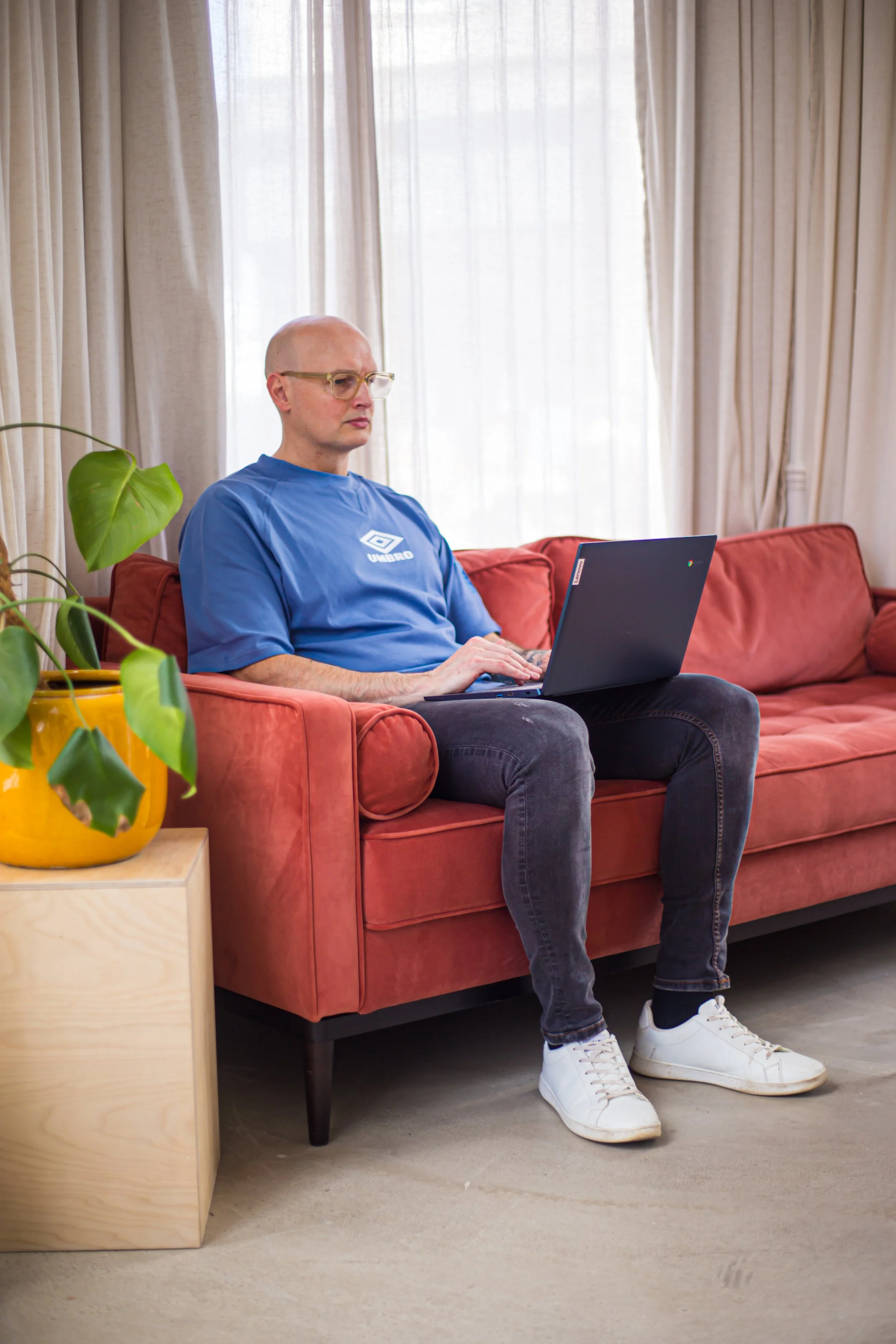 Lee Rober is sitting on a red sofa, using a laptop. He is wearing a blue Umbro t-shirt, black pants, and white sneakers. A green plant in a yellow pot is on a small wooden side table next to him, in front of beige curtains with light shining through.