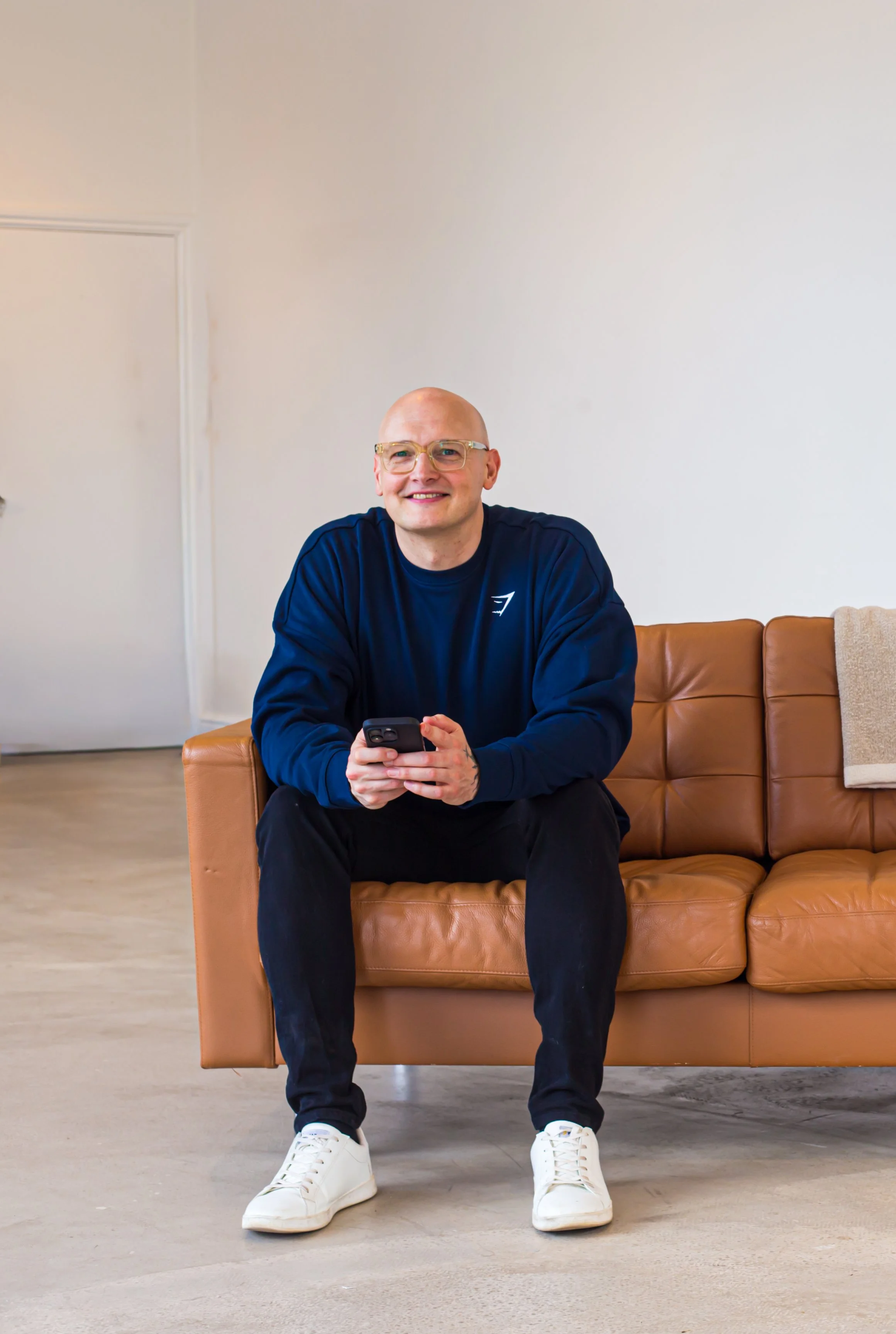 Lee Roberts is wearing glasses sitting on a tan leather sofa, wearing a navy blue sweatshirt, black pants, and white sneakers, holding a smartphone in his hands, in a room with a white wall and wooden floor.