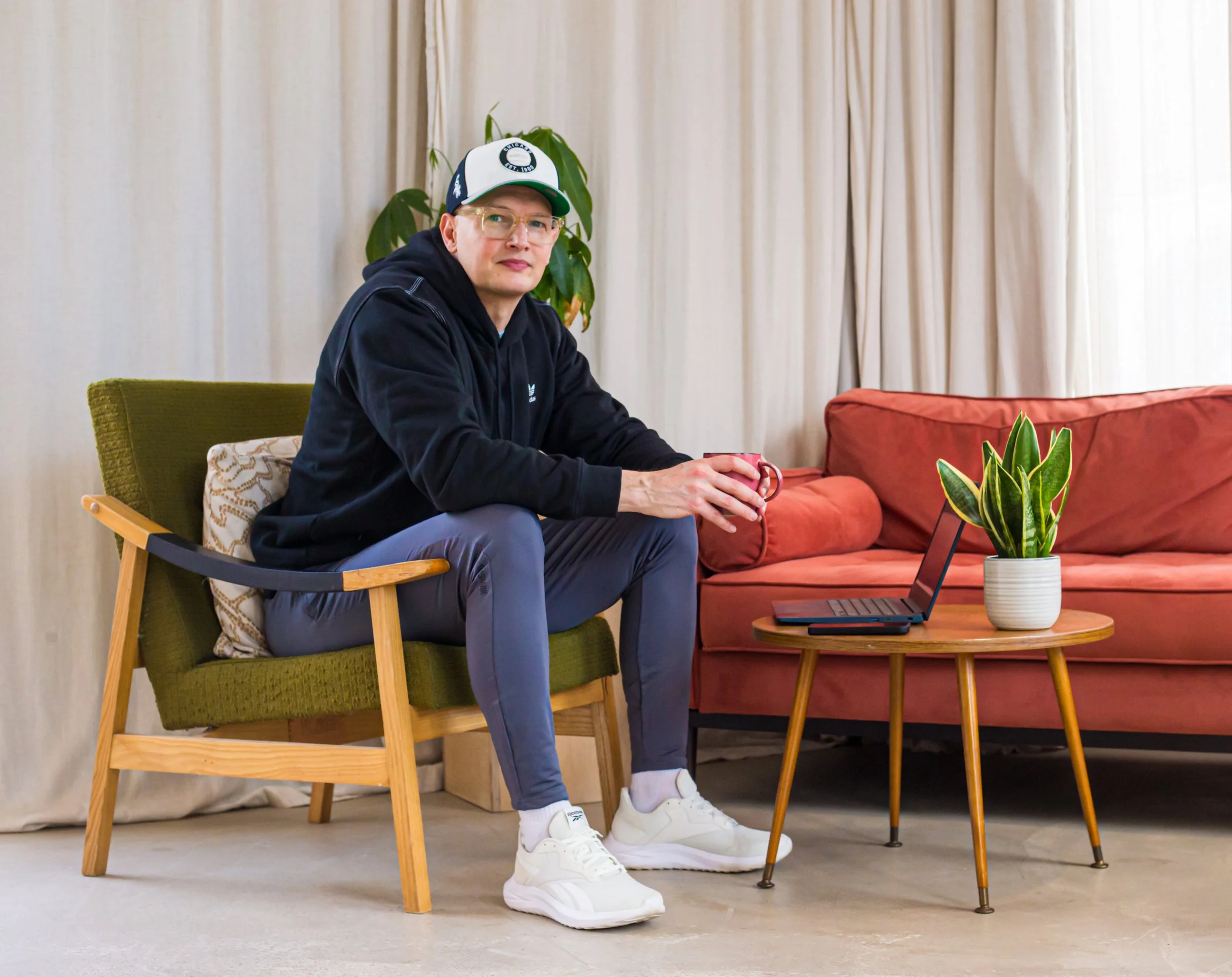 Lee Roberts sitting on a green armchair with wooden armrests, holding a red mug. Lee is seated at a coffee table with a laptop and a potted plant. Behind them are cream-colored curtains, a reddish-orange sofa, and a plant.