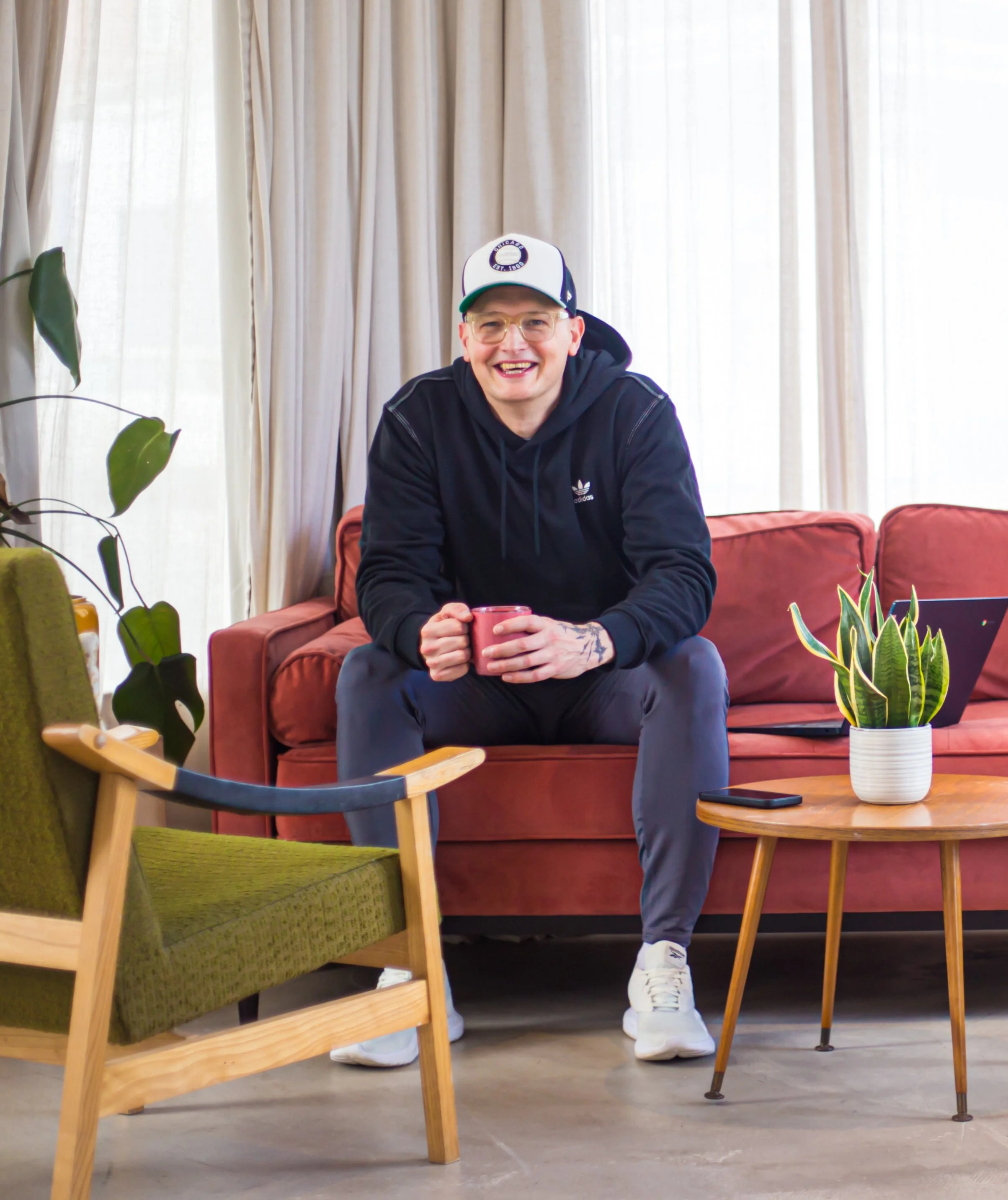Lee Roberts sitting on a couch holding a red mug, wearing glasses, a black hoodie, and white sneakers, with various chairs and plants in a room with beige curtains.