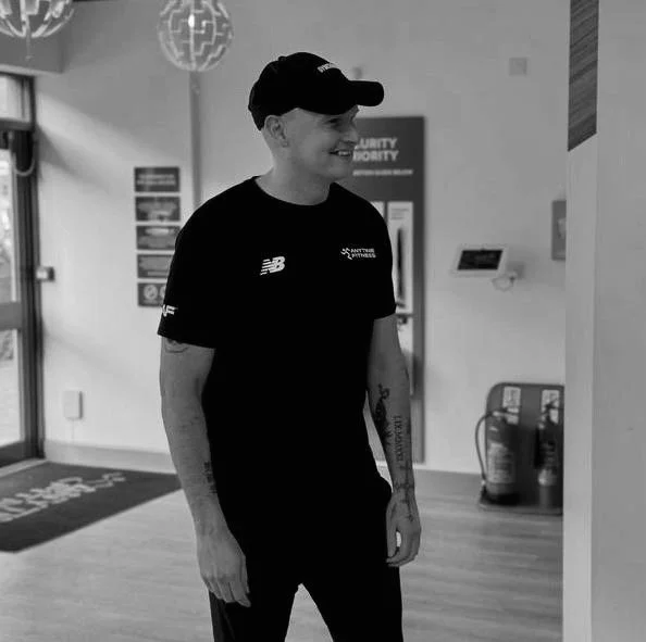 A young man smiling, wearing a black cap, black t-shirt, and black pants inside a gym or fitness center.