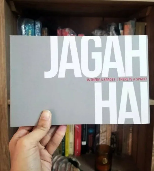 Person holding a book with large white and pink text on a gray background, in front of a wooden bookshelf filled with various books.