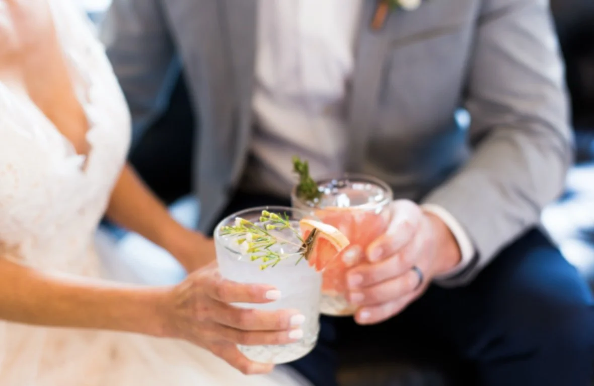Bride and Groom holding their signature cocktails with garnishes as they cheers for the wedding photographer.