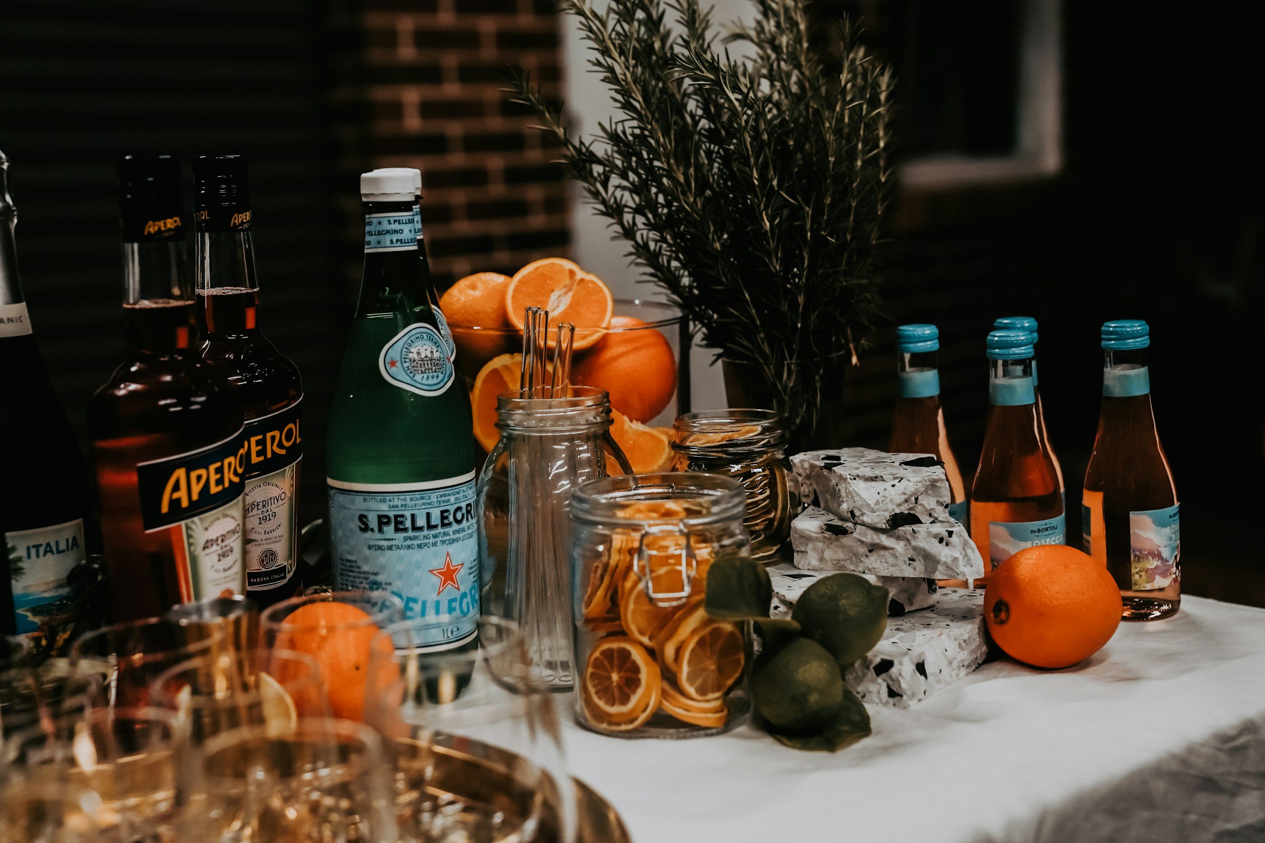 A mobile bar Aperol Spritz table set with bottles of Aperol and San Pellegrino, garnishes of lemon slices and limes, oranges, a bunch of rosemary, and small bottles of sparkling water, on a white tablecloth in a dimly lit setting.