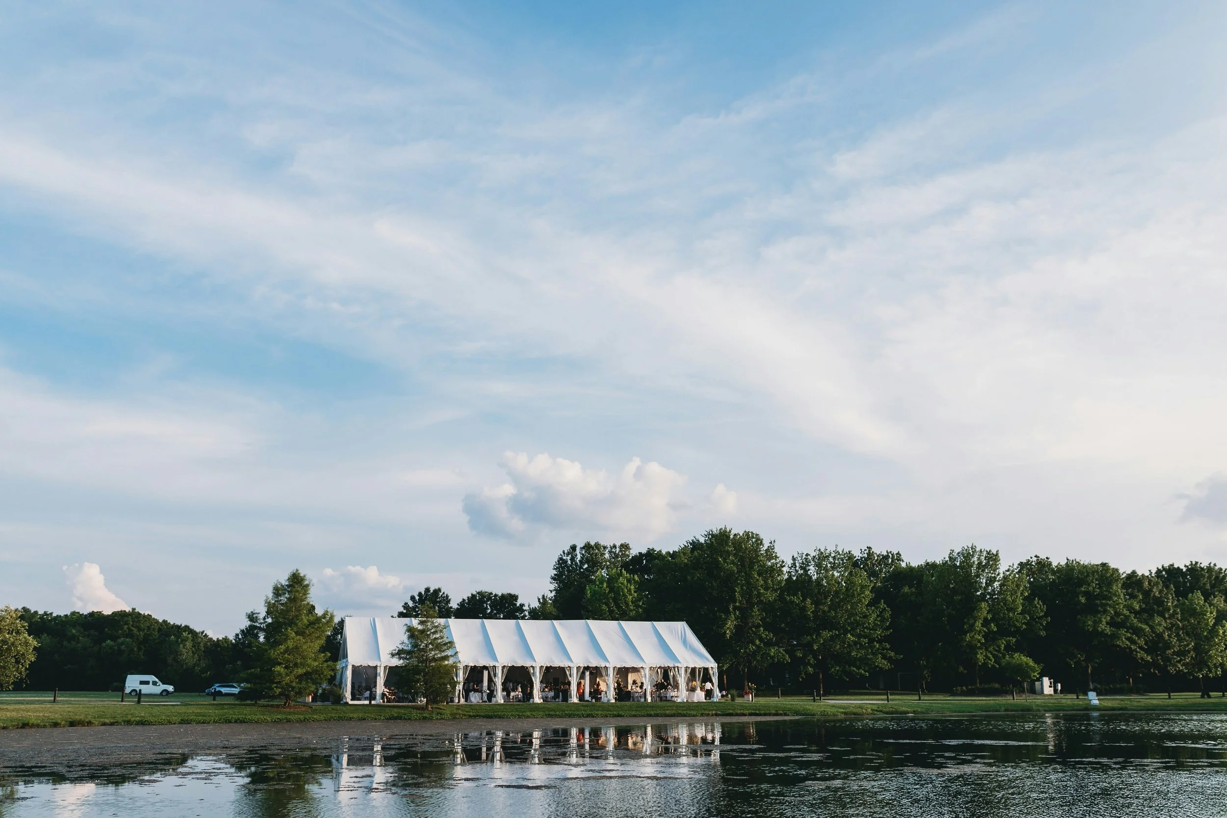 Large, white wedding tent set up on shore of Chesapeake Bay with trees, under a blue, partly cloudy sky.