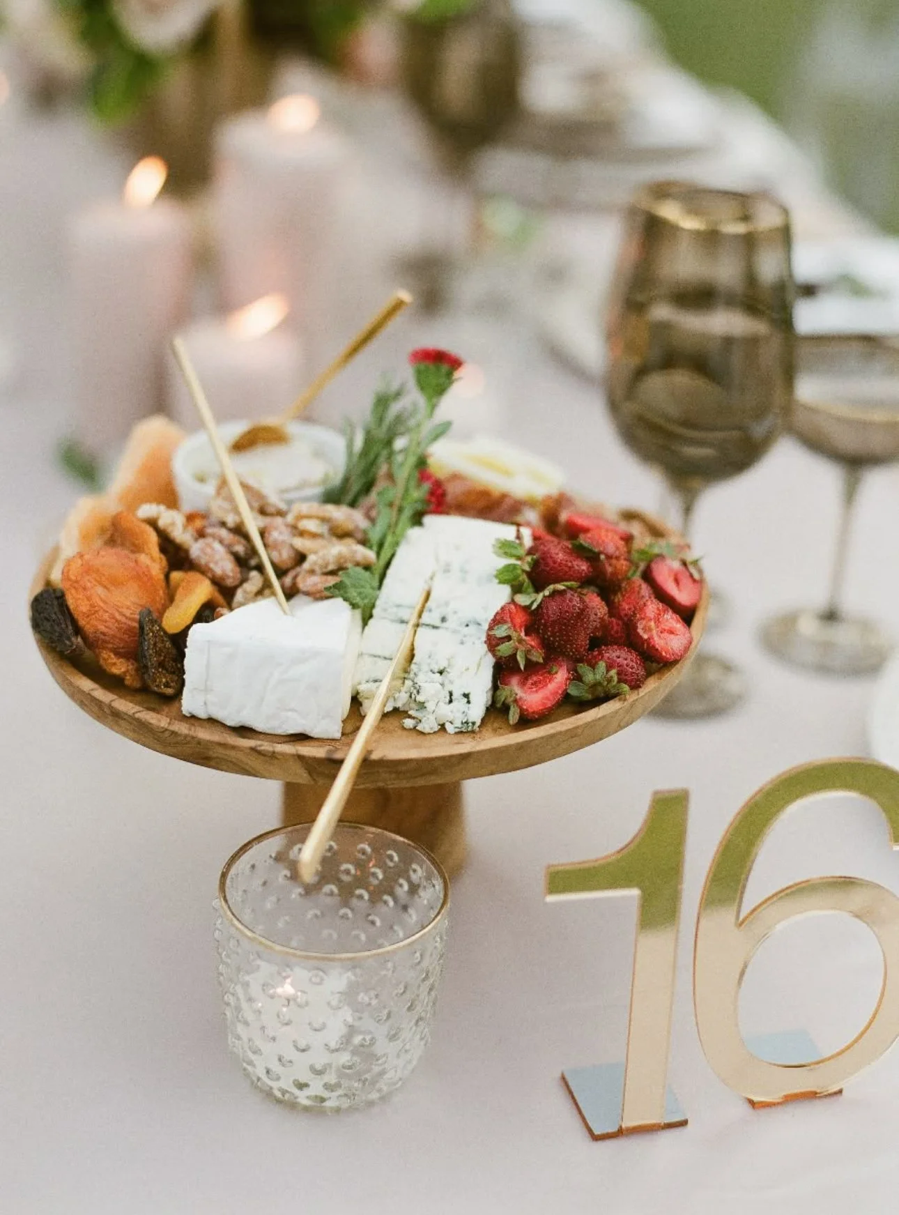 Cheese, strawberries, nuts, and dried fruits on a wooden platter at a celebration table with candlelight and drinks.