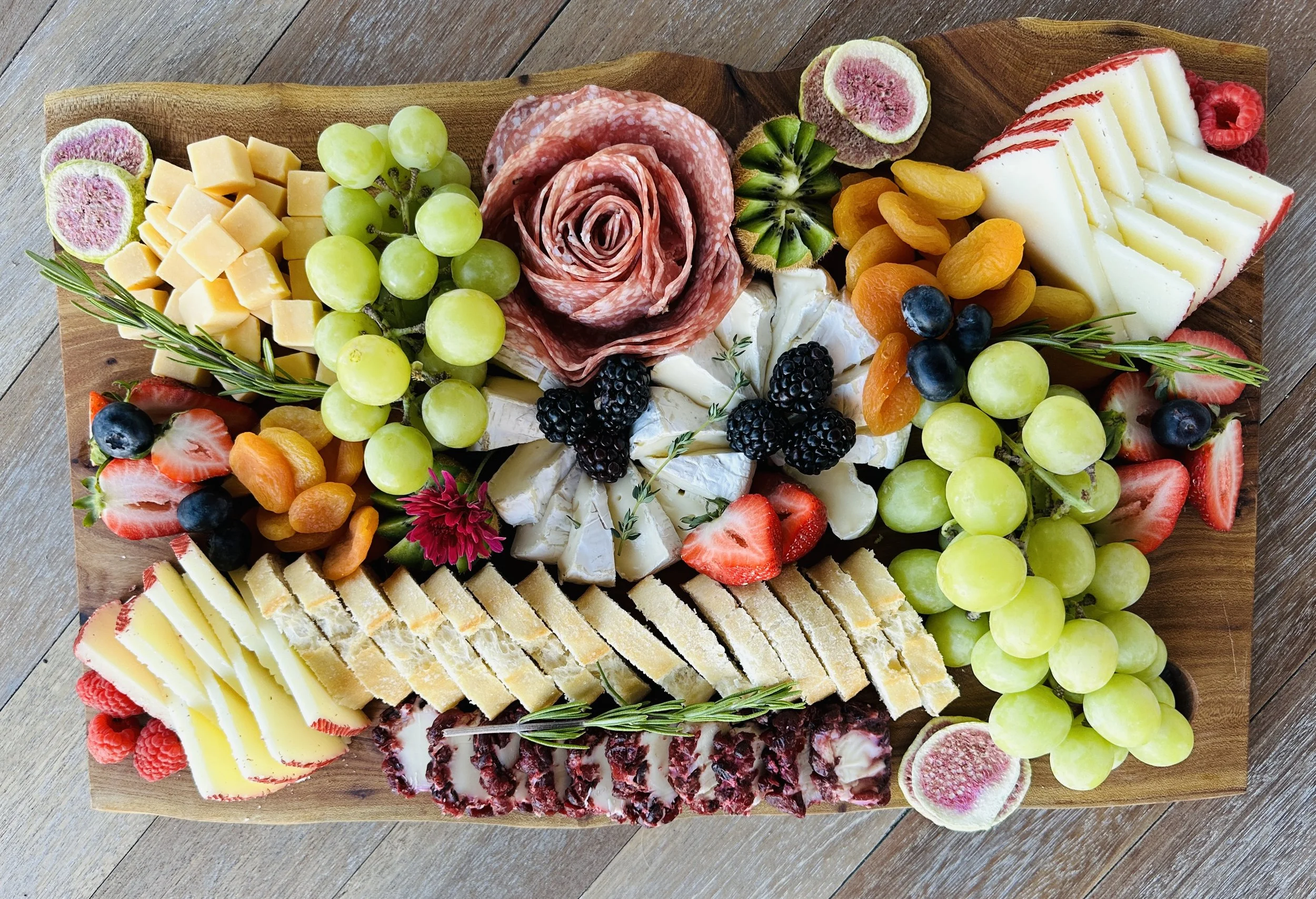 Charcuterie board graze with salami roses, decorative sliced cheeses, grapes, berries, dried apricots, sliced cured meats, and fresh garnishes like rosemary and edible flowers.