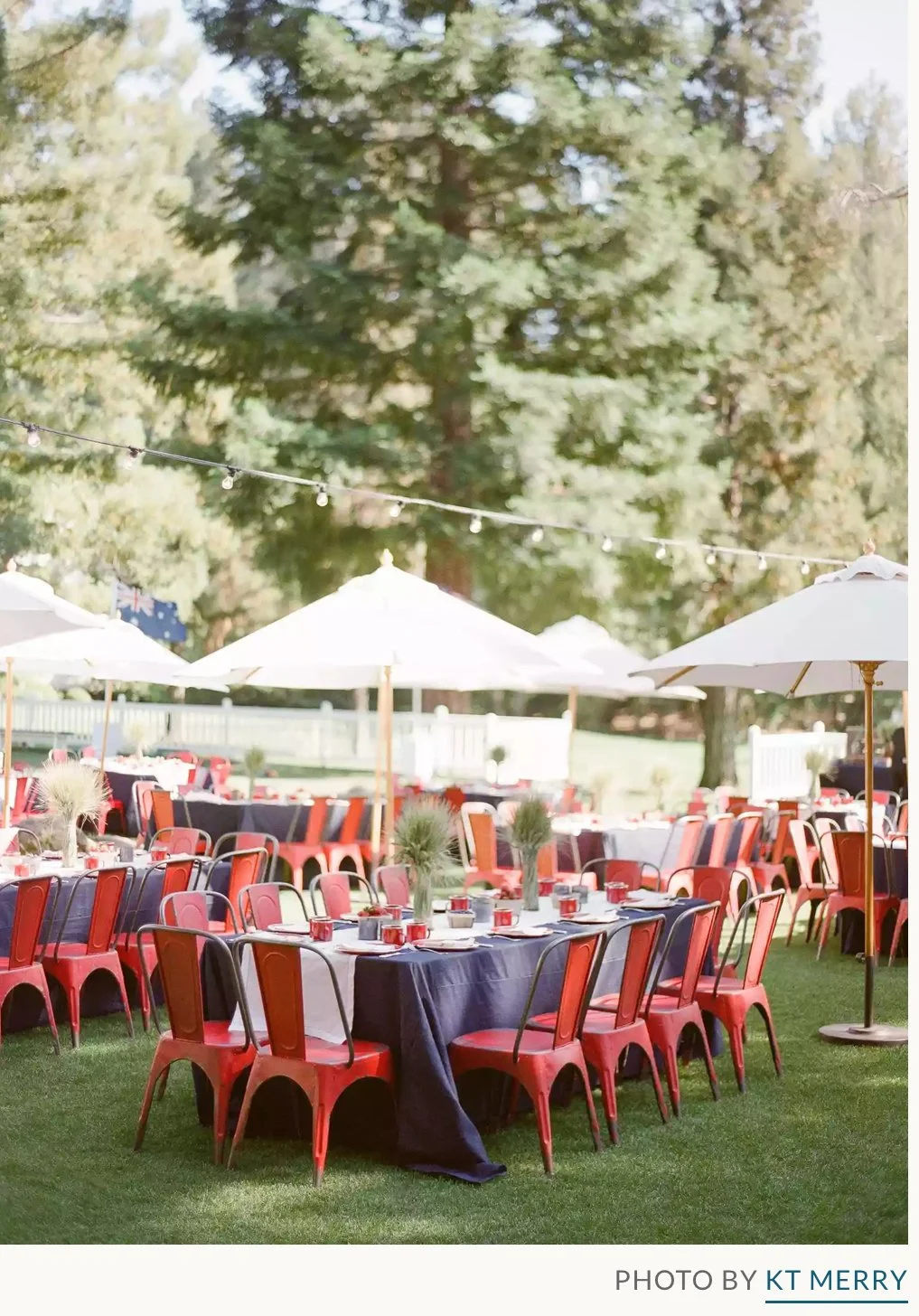 Outdoor holiday party event setup with round tables covered with navy blue tablecloths, red chairs, white umbrellas, and string lights against a backdrop of trees and a white fence.