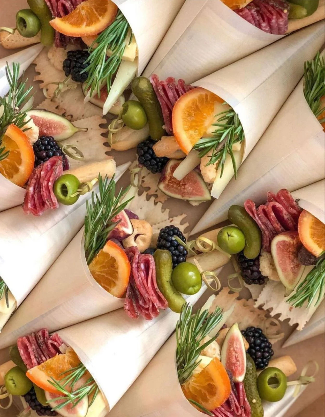 Charcuterie Cones wrapped in white paper with fresh artisan cheeses, cured meats, olives, fruit, nuts and crackers with rosemary sprig garnish. 